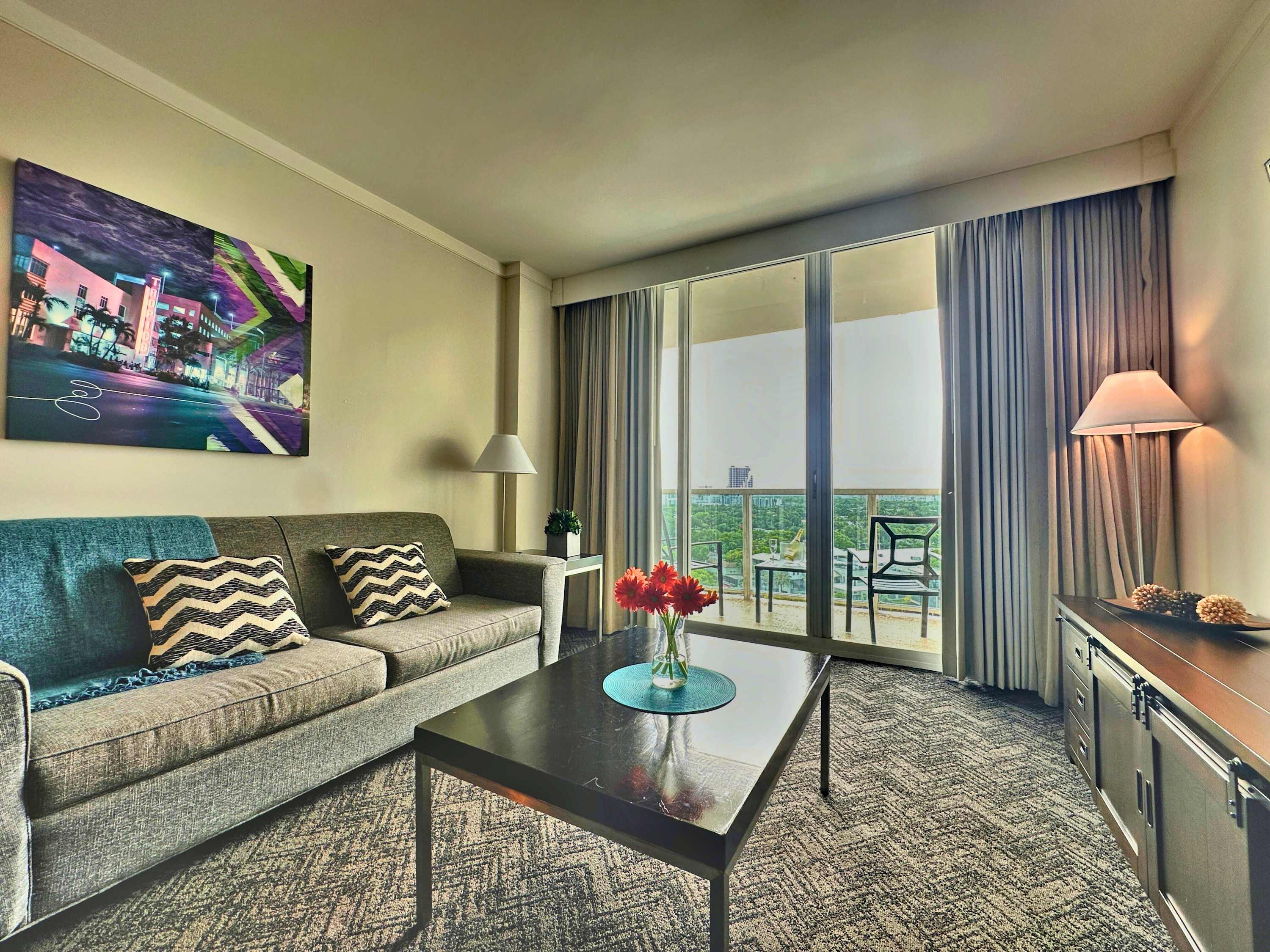 Living room with a gray sofa, patterned throw pillows, a dark coffee table, and floor-to-ceiling glass doors leading to a balcony with city views.