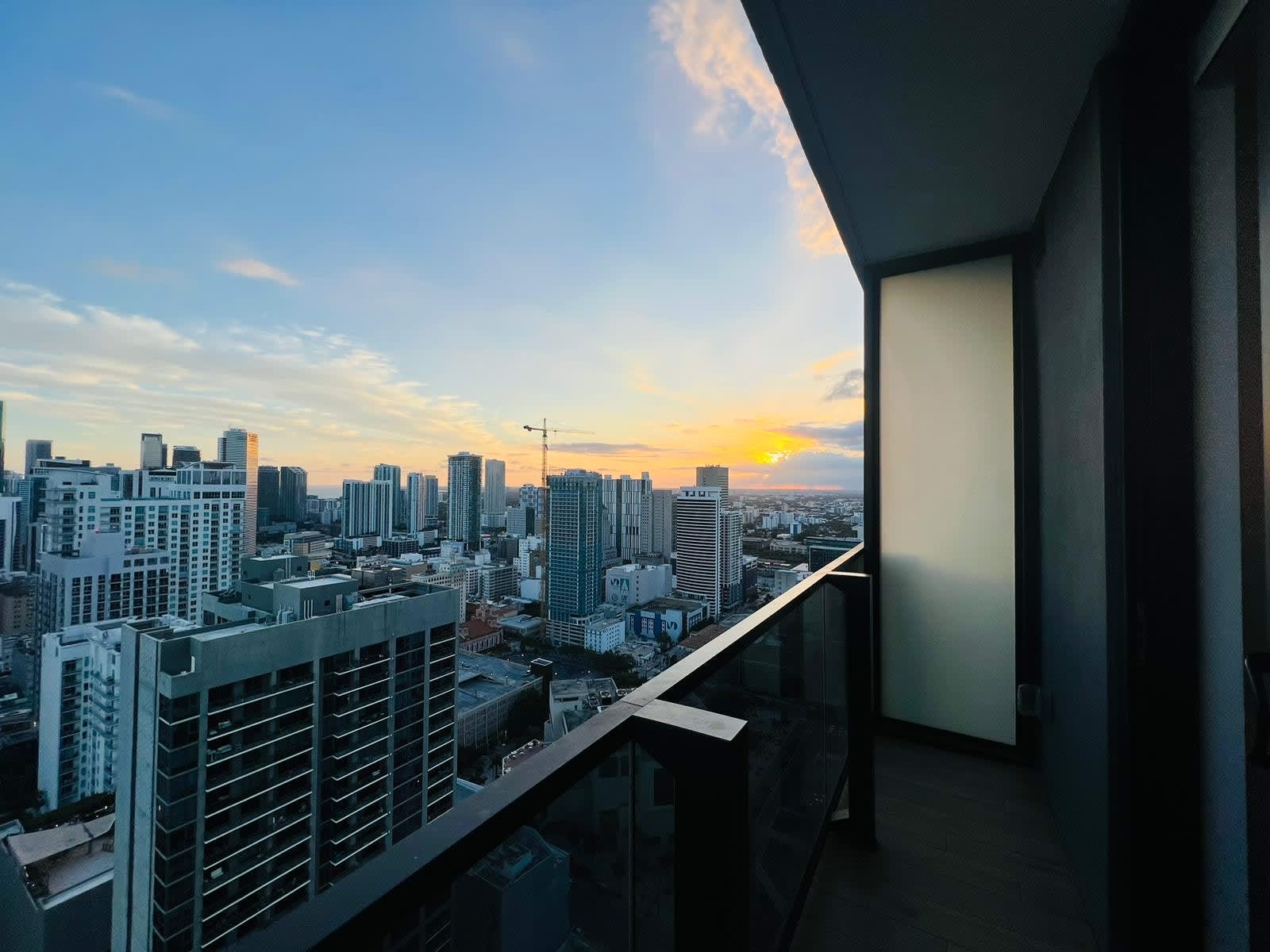 Balcony view of a sprawling city skyline bathed in the warm hues of a sunset, featuring a modern glass railing and dark wood-look flooring.