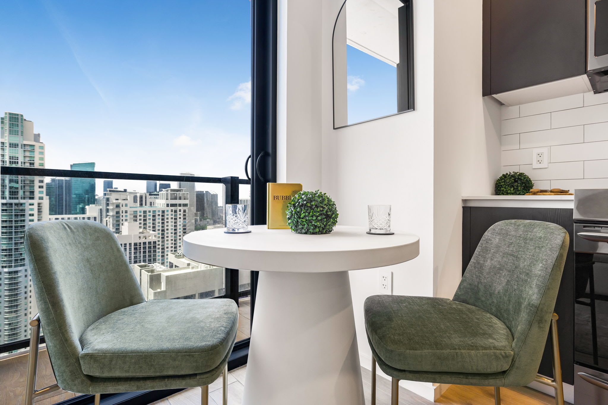 A compact dining area features a white round pedestal table with two green upholstered chairs, positioned next to a large window offering panoramic city views. A decorative mirror hangs on the wall, and a small plant adorns the table.