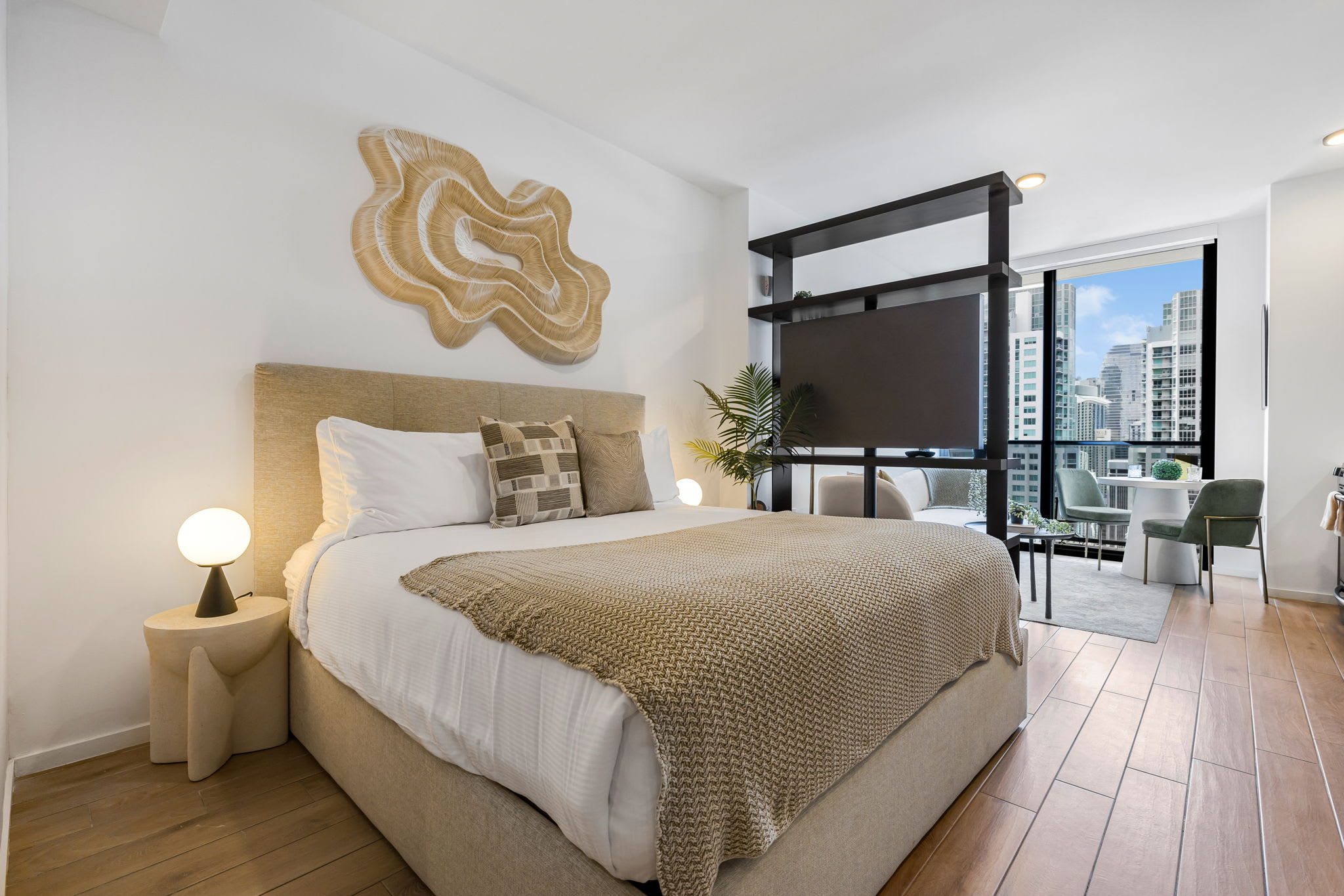 The studio bedroom features a queen-sized bed with a beige upholstered headboard and neutral bedding, complemented by a large abstract woven artwork on the white wall. A dark open shelving unit acts as a room divider, separating the sleeping area from the living space with city views.