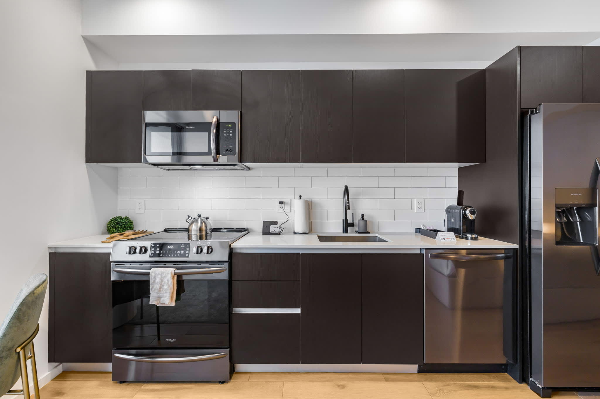 A sleek, modern kitchen features dark brown flat-panel cabinetry, stainless steel appliances including a microwave, range, and dishwasher, and a white subway tile backsplash. A light-colored countertop holds a sink with a black faucet and various kitchen items.