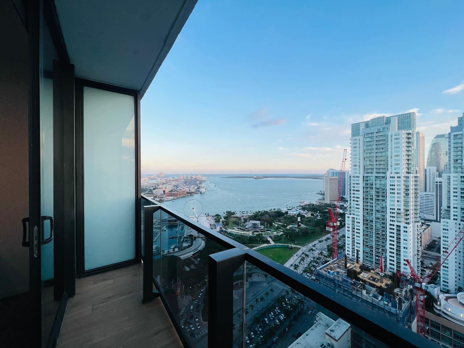 Balcony view showcasing a city skyline, a bay with a Ferris wheel, and distant buildings under a soft blue and pink sunset sky, featuring a modern glass railing and wood-look flooring.