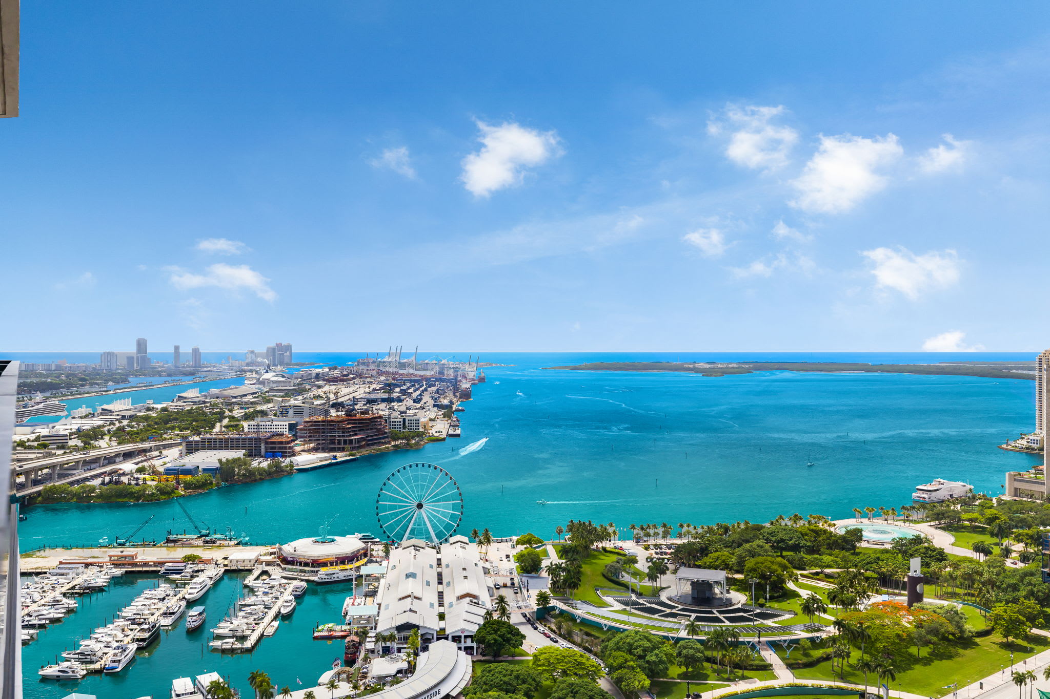 Panoramic aerial view of a vibrant coastal city with a large marina filled with boats, a prominent Ferris wheel, and numerous high-rise buildings along the waterfront, all under a clear blue sky.