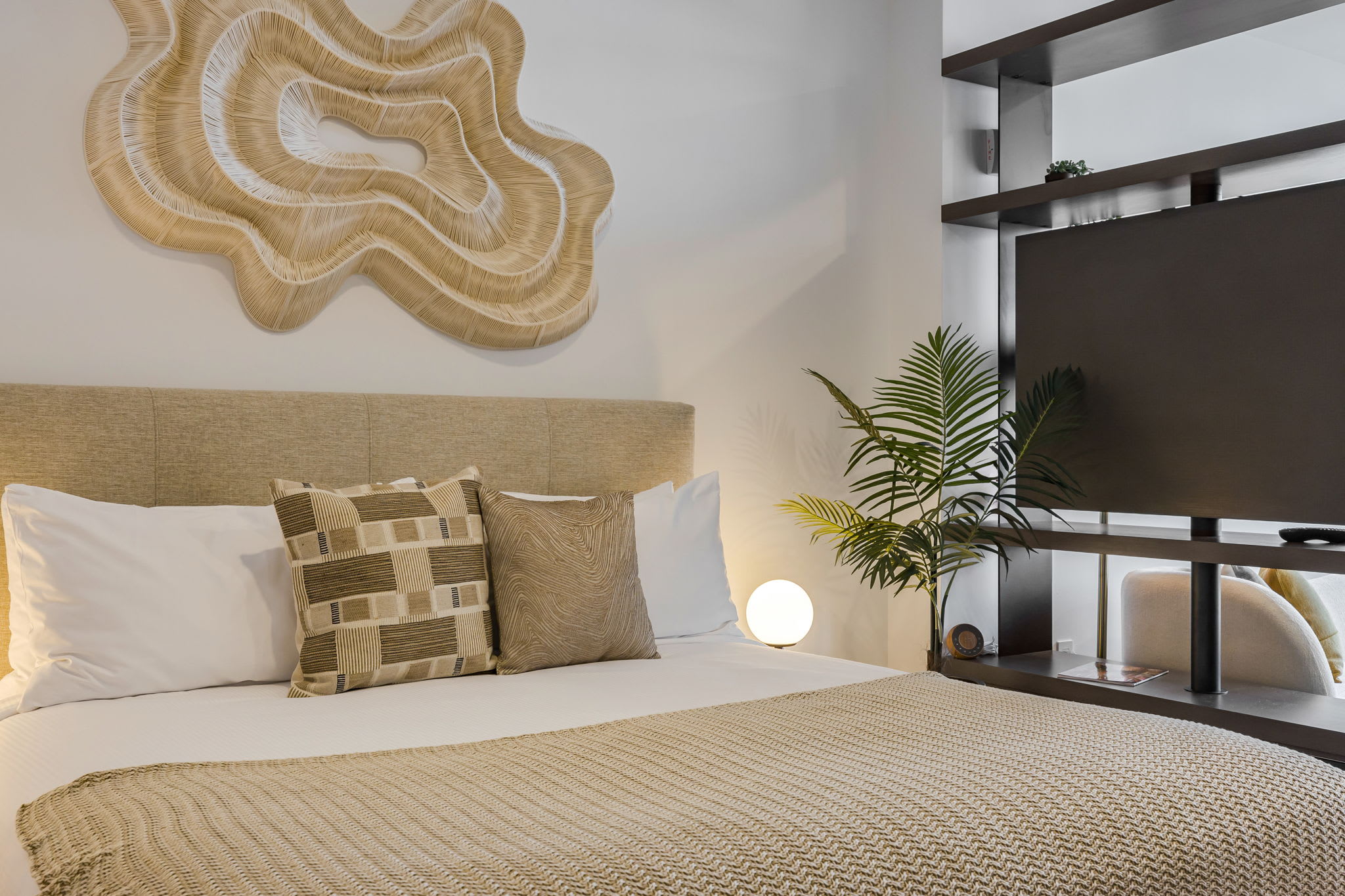 A close-up of the comfortable queen bed in the studio bedroom, featuring a beige upholstered headboard, white linens, and a textured neutral throw blanket. Accent pillows with geometric patterns add visual interest, while a modern table lamp sits on a unique sculptural nightstand.