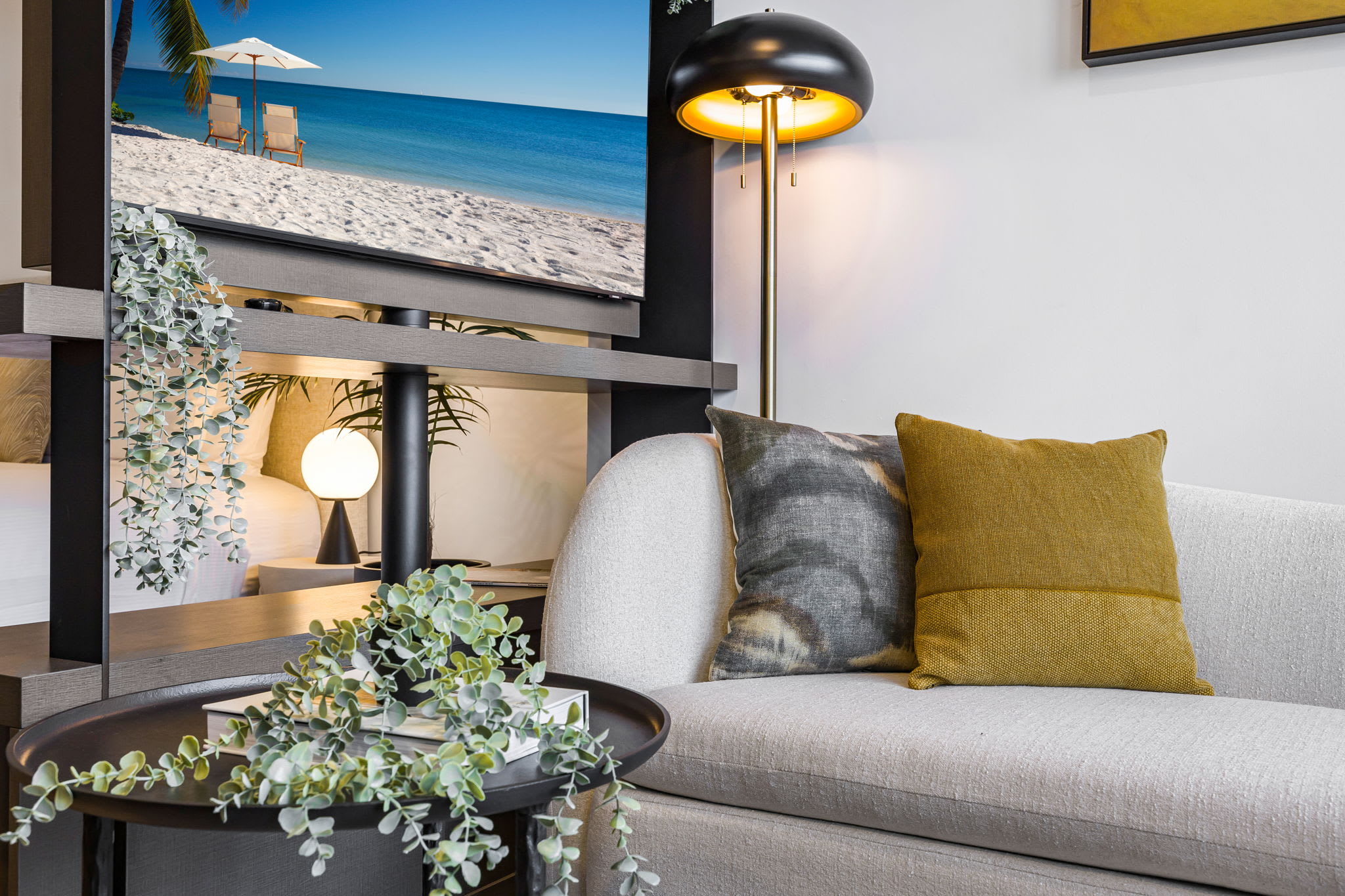 Detailed view of the living room's light-colored sofa with accent pillows, a dark round side table with greenery, and a stylish floor lamp, with a large TV screen visible in the background.