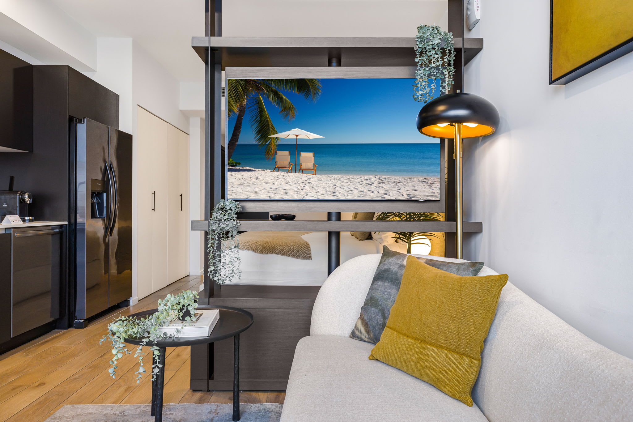 Cozy living space with a light-colored sofa, decorative pillows, and a dark side table, facing a large television integrated into a shelving unit, with a modern kitchen visible in the background.