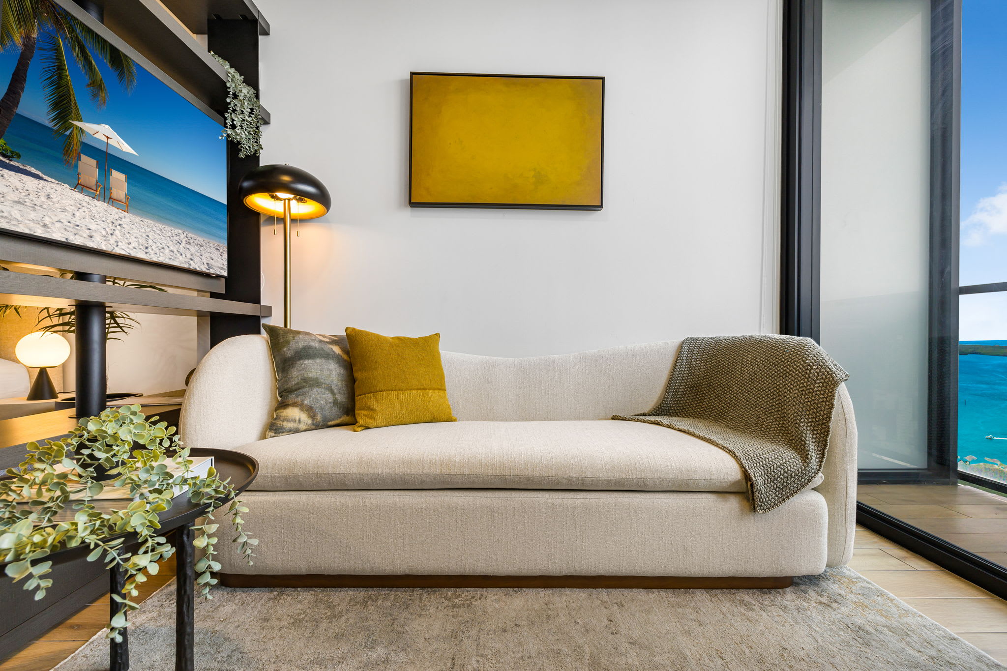 A modern living area features a light-colored curved sofa with accent pillows and a throw, a gold-framed artwork on a white wall, and a large window offering a stunning ocean view. A multi-tiered shelf with a large screen displaying a beach scene adds a unique touch.