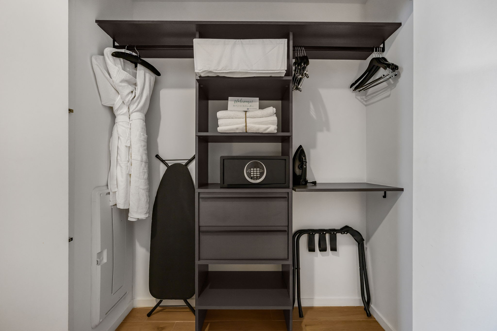 Organized closet space featuring dark shelving with folded towels, a safe, an iron, an ironing board, and a white bathrobe hanging, providing essential guest amenities.