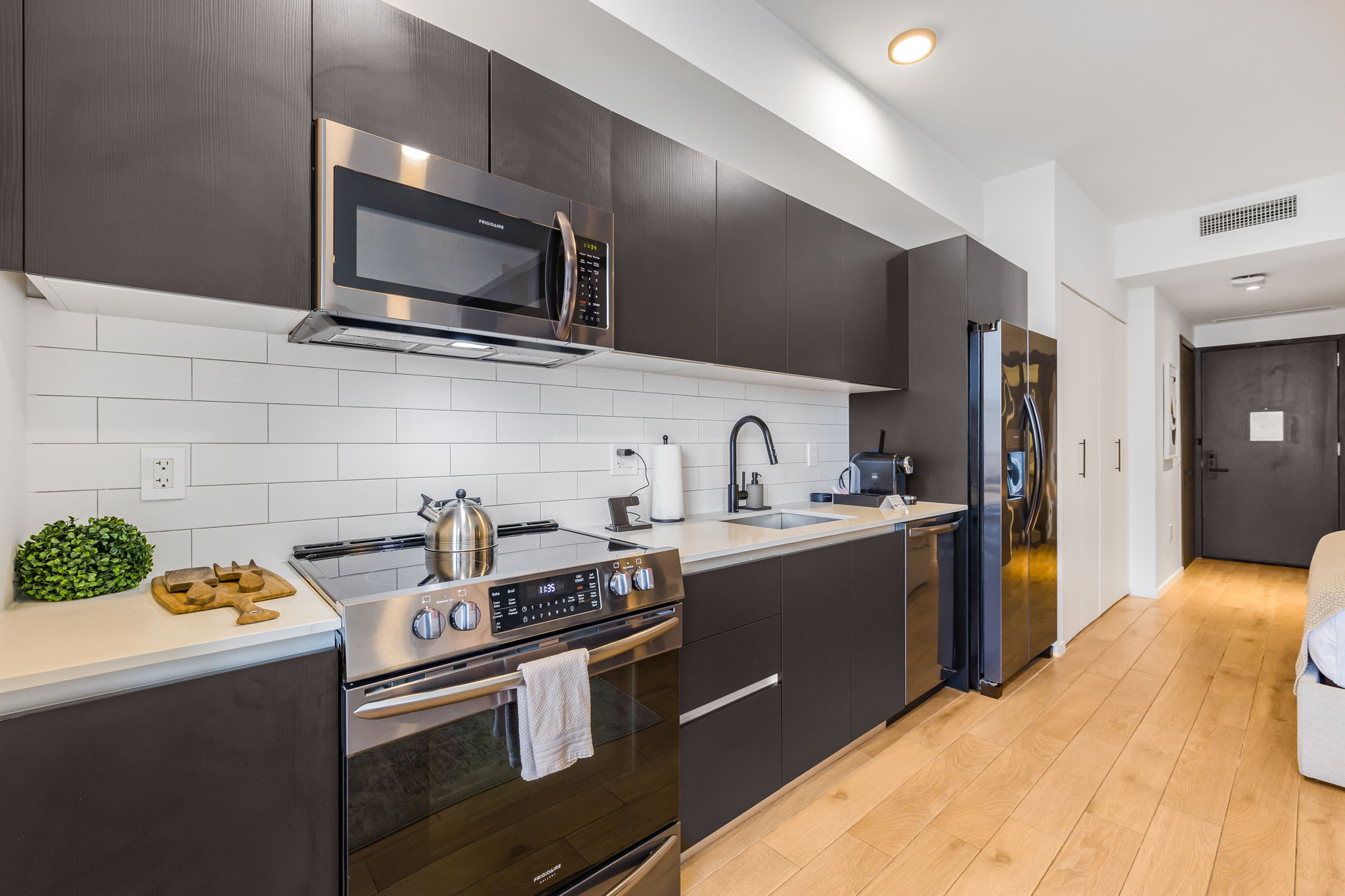 A well-appointed kitchen showcases dark brown cabinetry, stainless steel appliances including a microwave, electric range, and a large refrigerator, set against a white subway tile backsplash. Light hardwood floors extend throughout the space, leading towards the main living area.