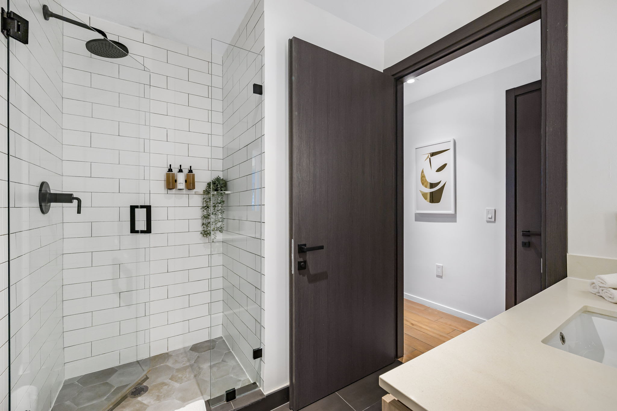 A spacious walk-in shower features white subway tiles on the walls and hexagonal grey floor tiles, enclosed by a clear glass panel. Black matte fixtures, including a rain showerhead and a handheld shower, provide a contemporary look, with small plants adding a decorative touch.