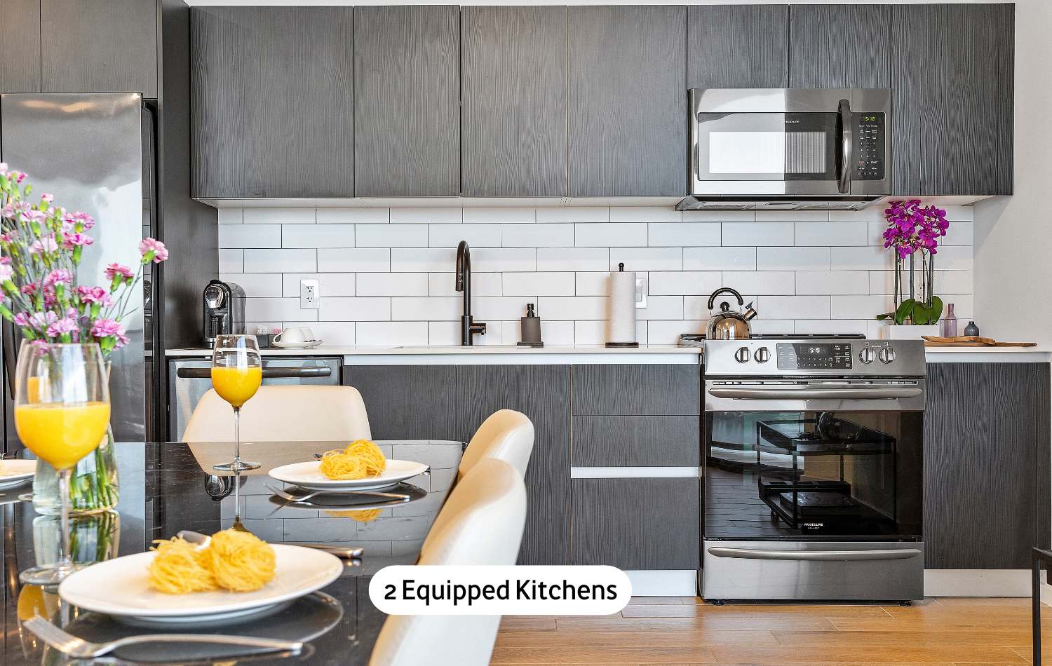 A stylish, fully equipped kitchen featuring dark wood-grain cabinetry, a white subway tile backsplash, stainless steel appliances, and a black countertop, with a dining table set for a meal in the foreground.