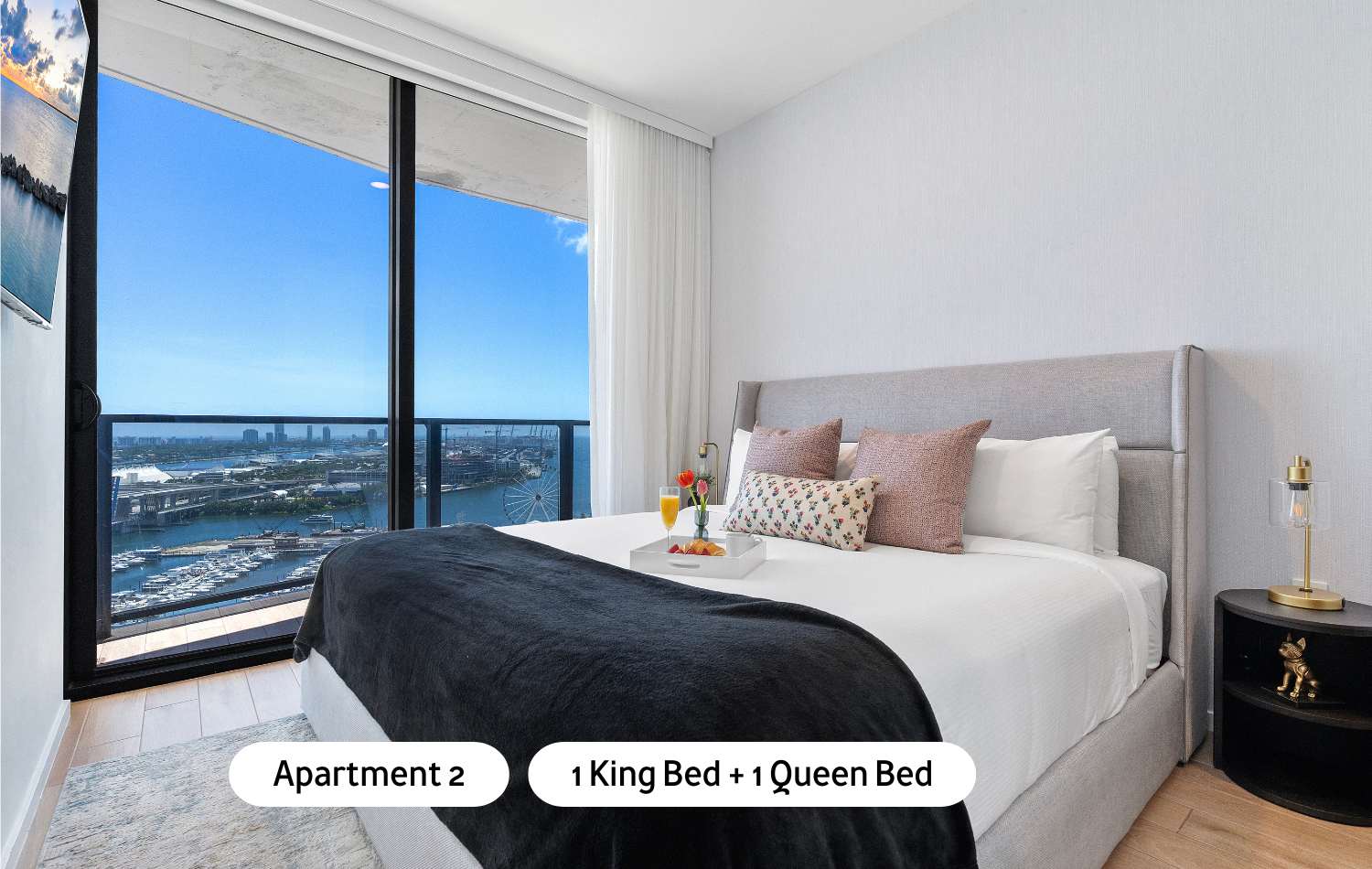 A second elegant bedroom featuring a king-sized bed with a light grey upholstered headboard, a nightstand, and large floor-to-ceiling windows providing a breathtaking ocean and city view.