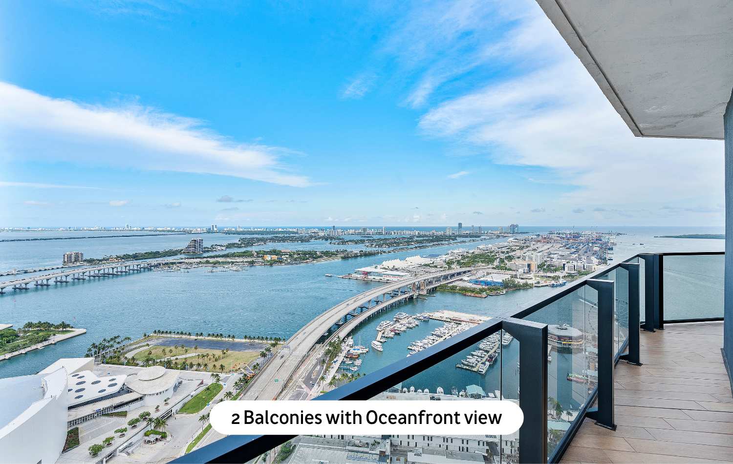 An expansive balcony with a clear glass railing, providing a breathtaking panoramic oceanfront view of the city skyline, marina, and distant bridges under a bright blue sky.