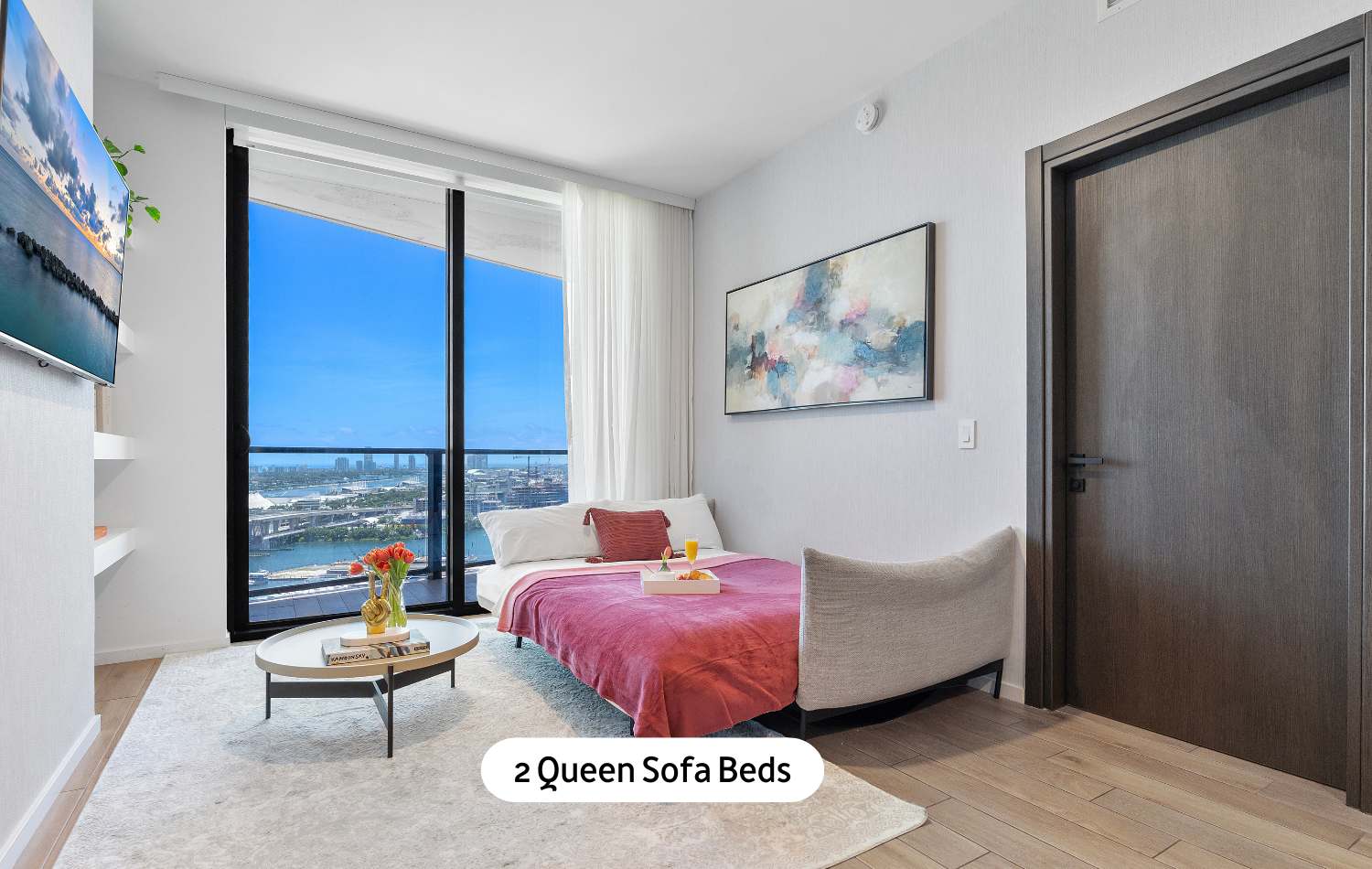 A versatile room featuring a queen sofa bed with a pink throw, a round coffee table, and expansive floor-to-ceiling windows offering panoramic ocean and city views, with a wall-mounted TV.