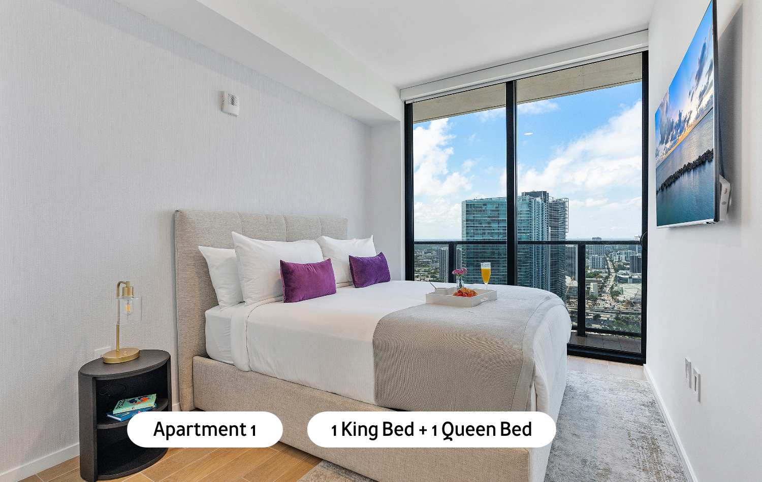A bright and modern bedroom featuring a king-sized bed with a light grey upholstered headboard, two nightstands, and expansive floor-to-ceiling windows offering a stunning city skyline view.