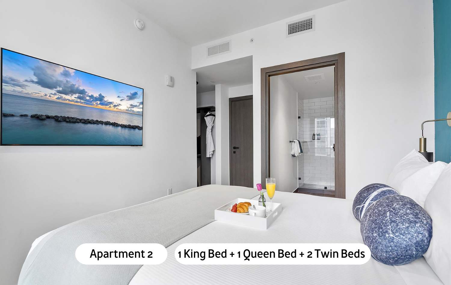 Another spacious bedroom with a king-sized bed, white bedding, a large wall-mounted television, and a glimpse of an en-suite bathroom entrance, featuring light walls and a modern aesthetic.