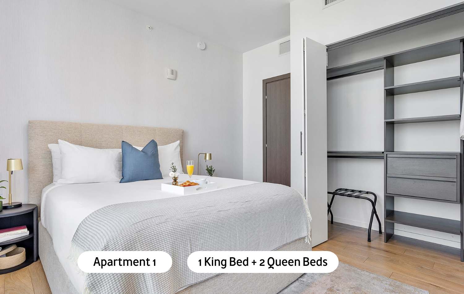 A bright bedroom featuring a king-sized bed with a light upholstered headboard, crisp white bedding, and a built-in dark wood open closet system with shelves and drawers, complemented by light walls and a rug.