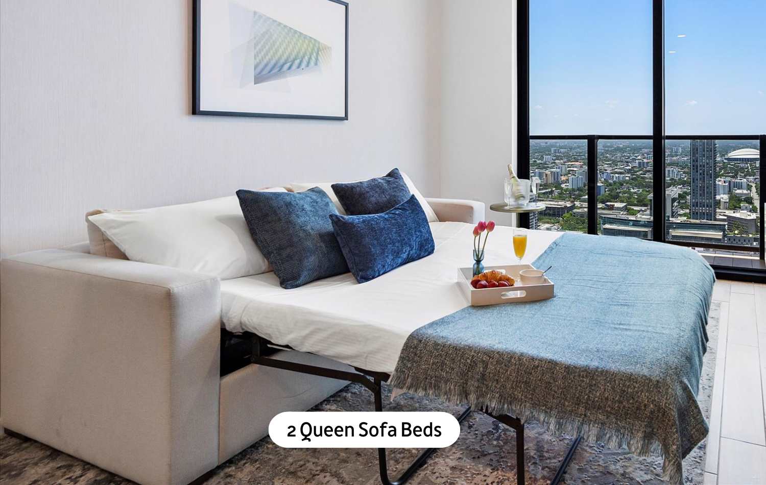 A versatile living space featuring a queen sofa bed pulled out, adorned with blue pillows and a throw blanket, positioned near a large window offering city views, on a light-colored rug.