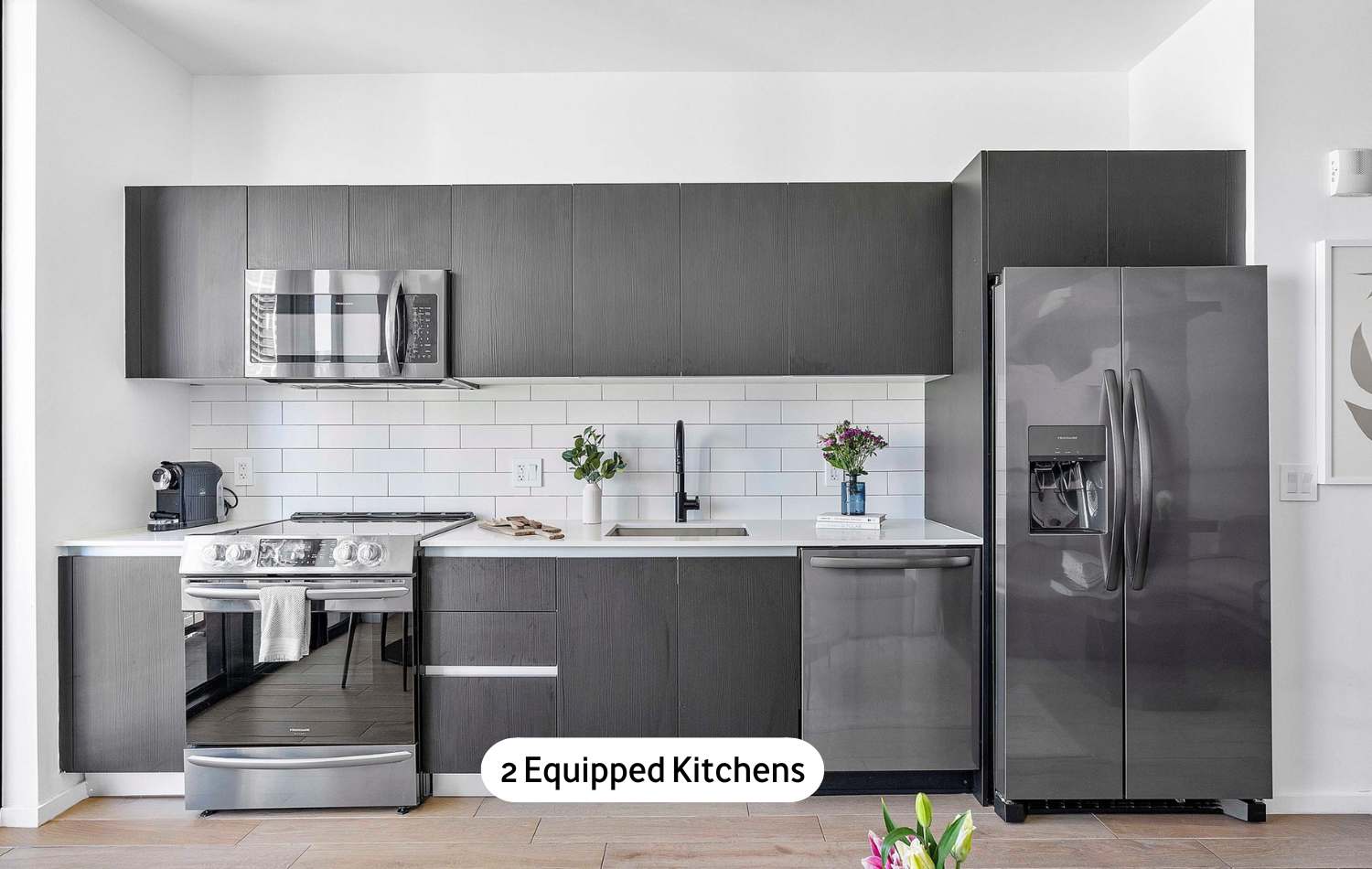 A sleek, modern kitchen featuring dark wood-grain cabinetry, white subway tile backsplash, and a suite of stainless steel appliances including a refrigerator, oven, microwave, and dishwasher, all set on light wood flooring.