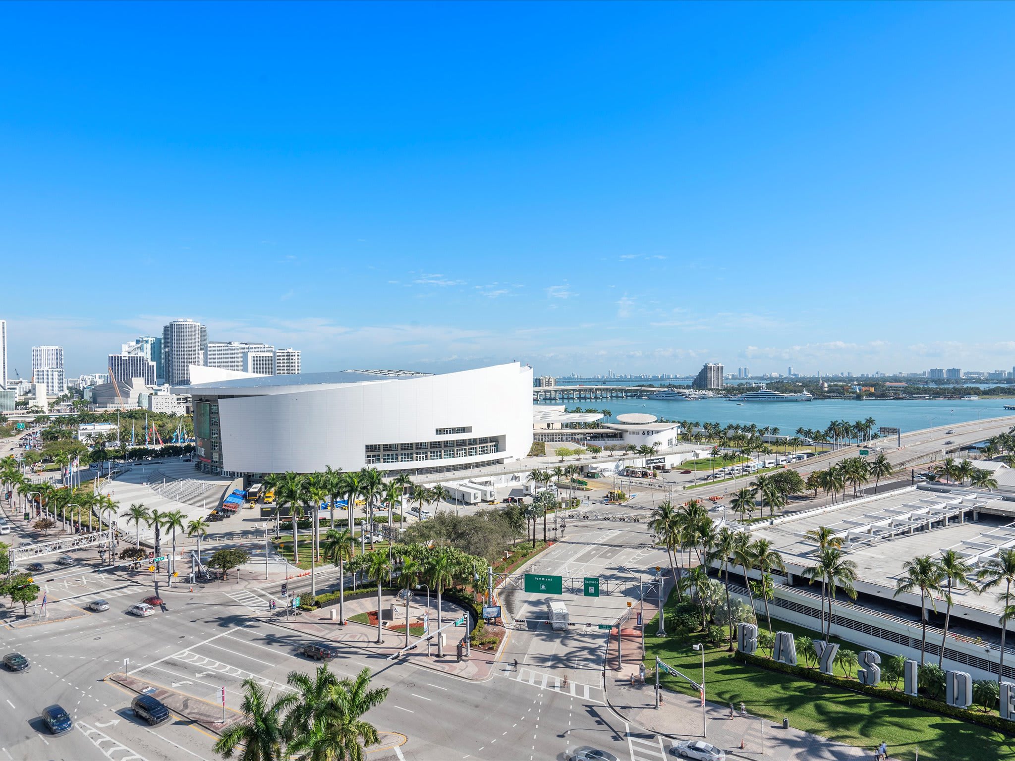 A wide aerial view of a bustling city with a prominent white stadium building, multiple roads with traffic, palm trees, and a large body of water with bridges and boats in the background under a bright blue sky.