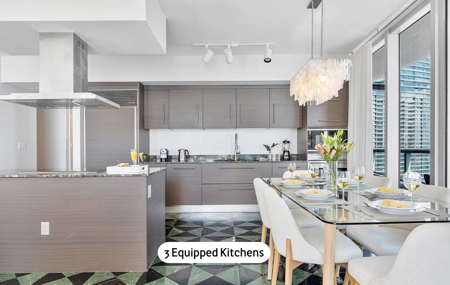 A sleek, modern kitchen featuring gray cabinetry, stainless steel appliances, a central island, and an adjacent dining area with a glass table and white chairs, all illuminated by a striking chandelier and natural light from large windows.