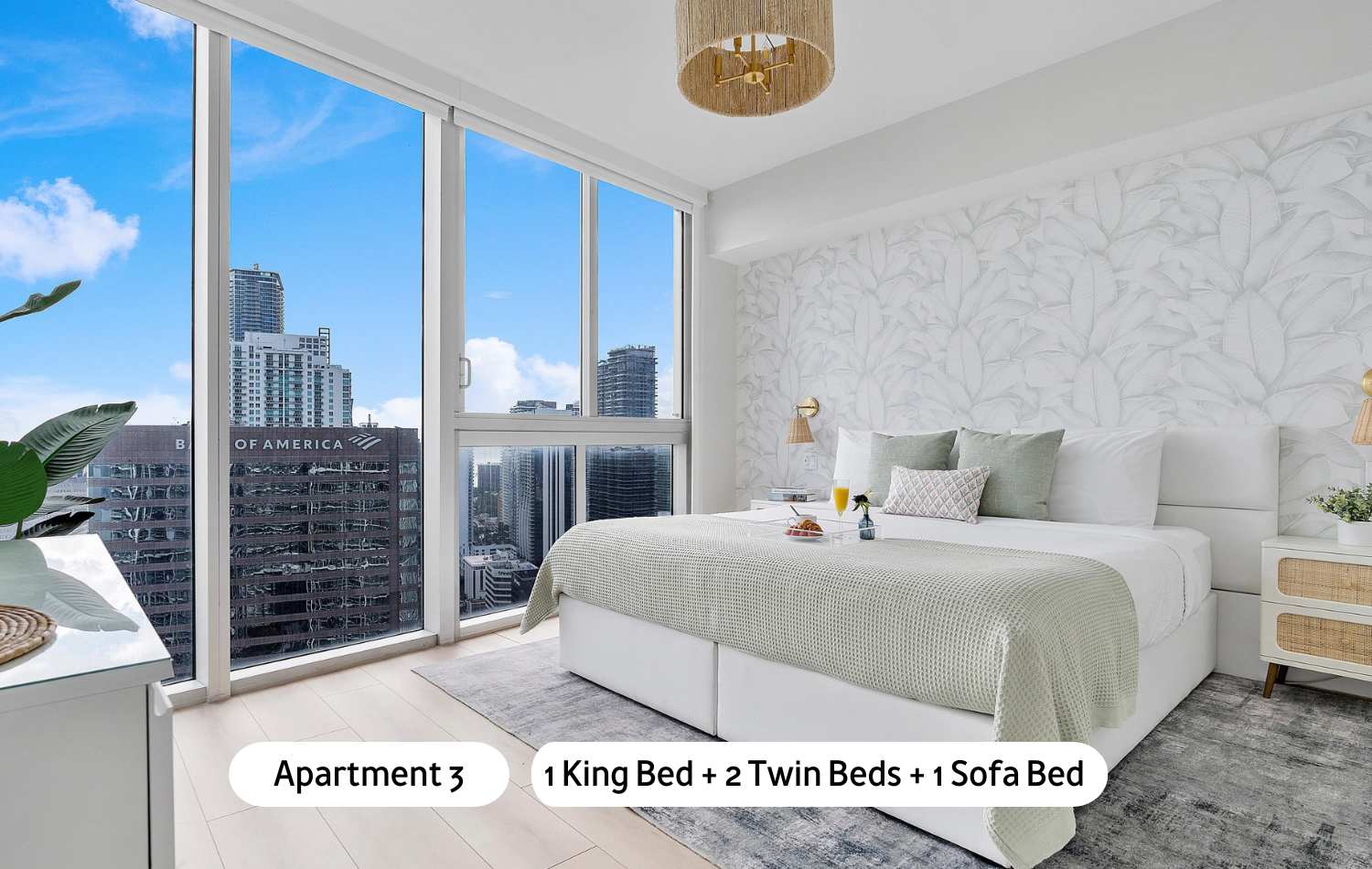 A spacious bedroom featuring a king-sized bed with light green bedding and two twin beds, complemented by light wood floors and a patterned rug, with expansive windows offering panoramic city views.