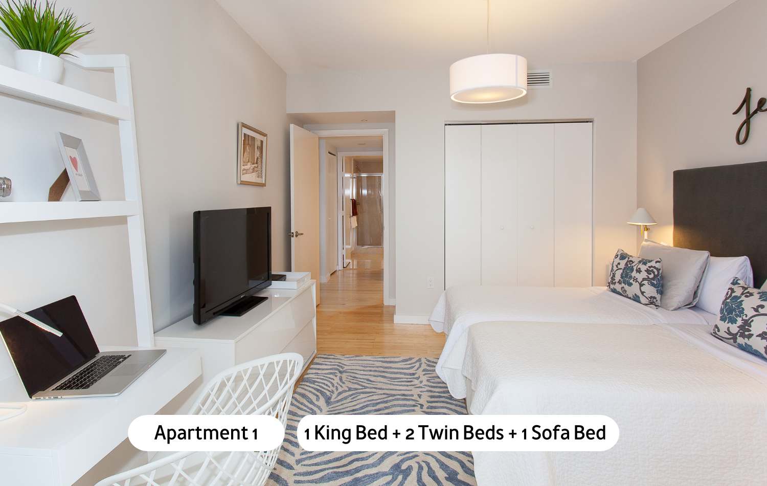 A bright bedroom furnished with two twin beds, a white desk and chair, a wall-mounted TV, and a white shelving unit, featuring light walls and a blue patterned rug, with a doorway leading to an interior hallway.
