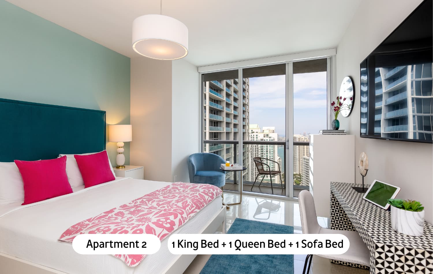 A vibrant bedroom featuring a king-sized bed with a blue headboard and pink accents, a stylish patterned desk, and a blue armchair, all set against large windows that provide city views and access to a private balcony.