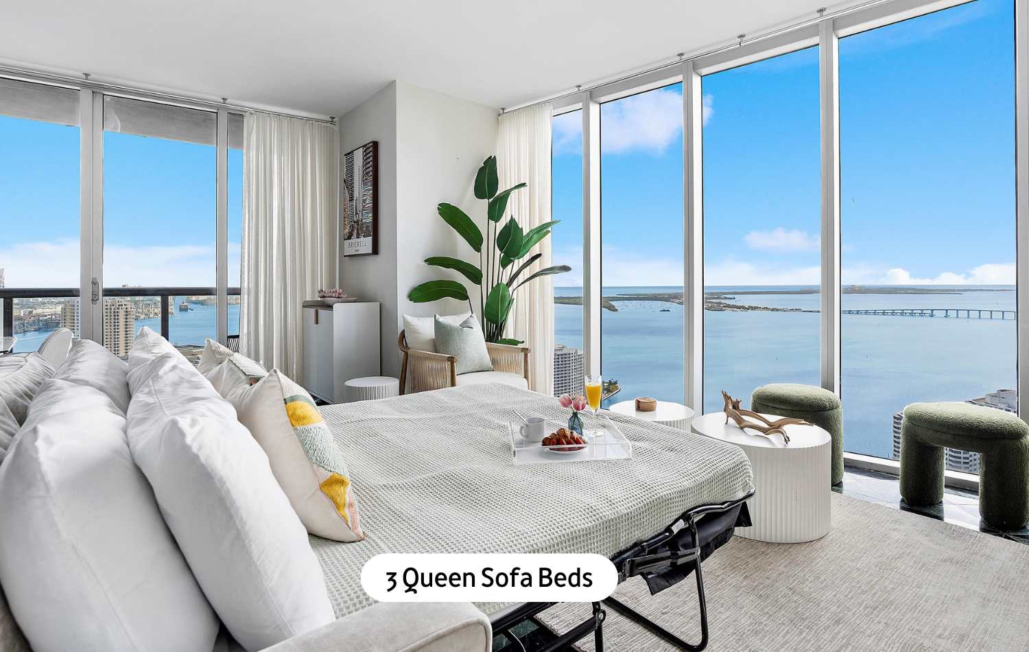 A bright living area featuring a comfortable sofa bed, a large potted plant, and floor-to-ceiling windows that offer breathtaking river and sea views, with a small table holding a breakfast tray.