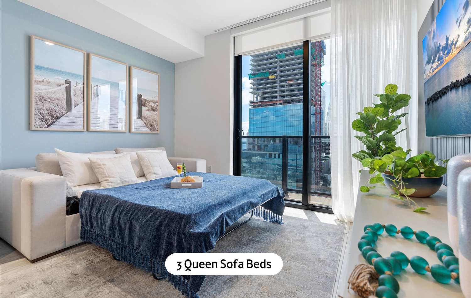 Living room transformed into a sleeping area with a queen sofa bed pulled out, covered with a blue throw blanket. A round coffee table sits in front, and a large window offers a city view.