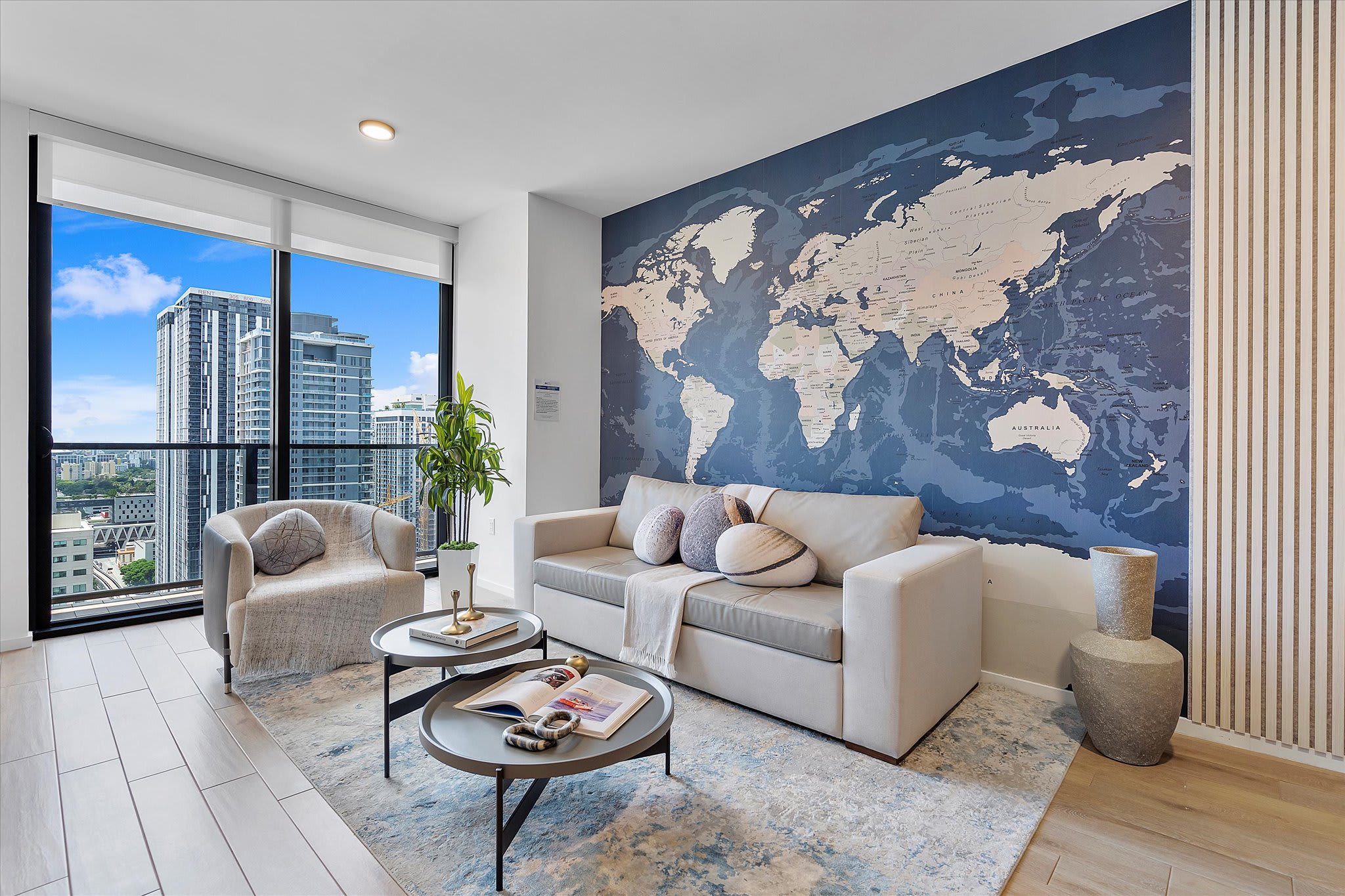 Contemporary living room with a large blue world map accent wall and light wood-look tile flooring. A light gray sofa with decorative pillows and a matching armchair are arranged around two round coffee tables, with floor-to-ceiling windows providing city views.