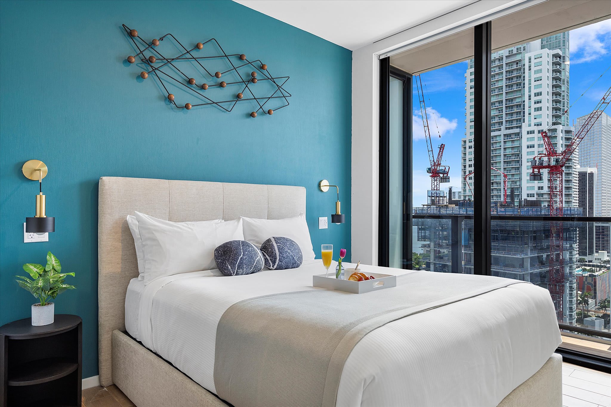 Stylish bedroom featuring a king-sized bed with a beige upholstered headboard, a teal accent wall with geometric metal artwork, and floor-to-ceiling windows offering city views. A breakfast tray is on the bed.