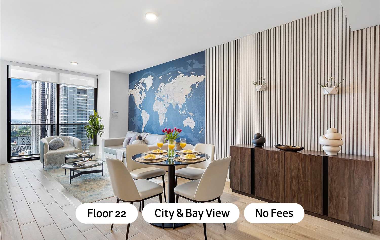 Modern open-concept living and dining area with light wood-look tile floors, a large blue world map accent wall, and floor-to-ceiling windows offering city views. A round glass dining table with four chairs is set for a meal, and a light gray sofa and armchair furnish the living space.