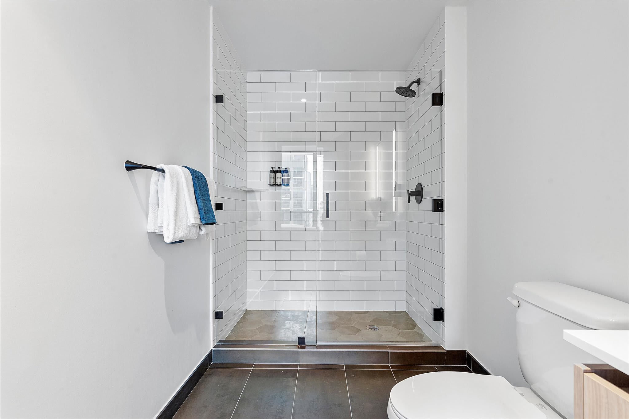 Contemporary bathroom featuring a spacious walk-in shower with white subway tiles and a glass enclosure, black fixtures, and a white toilet. The floor has dark large tiles and hexagon shower floor tiles.