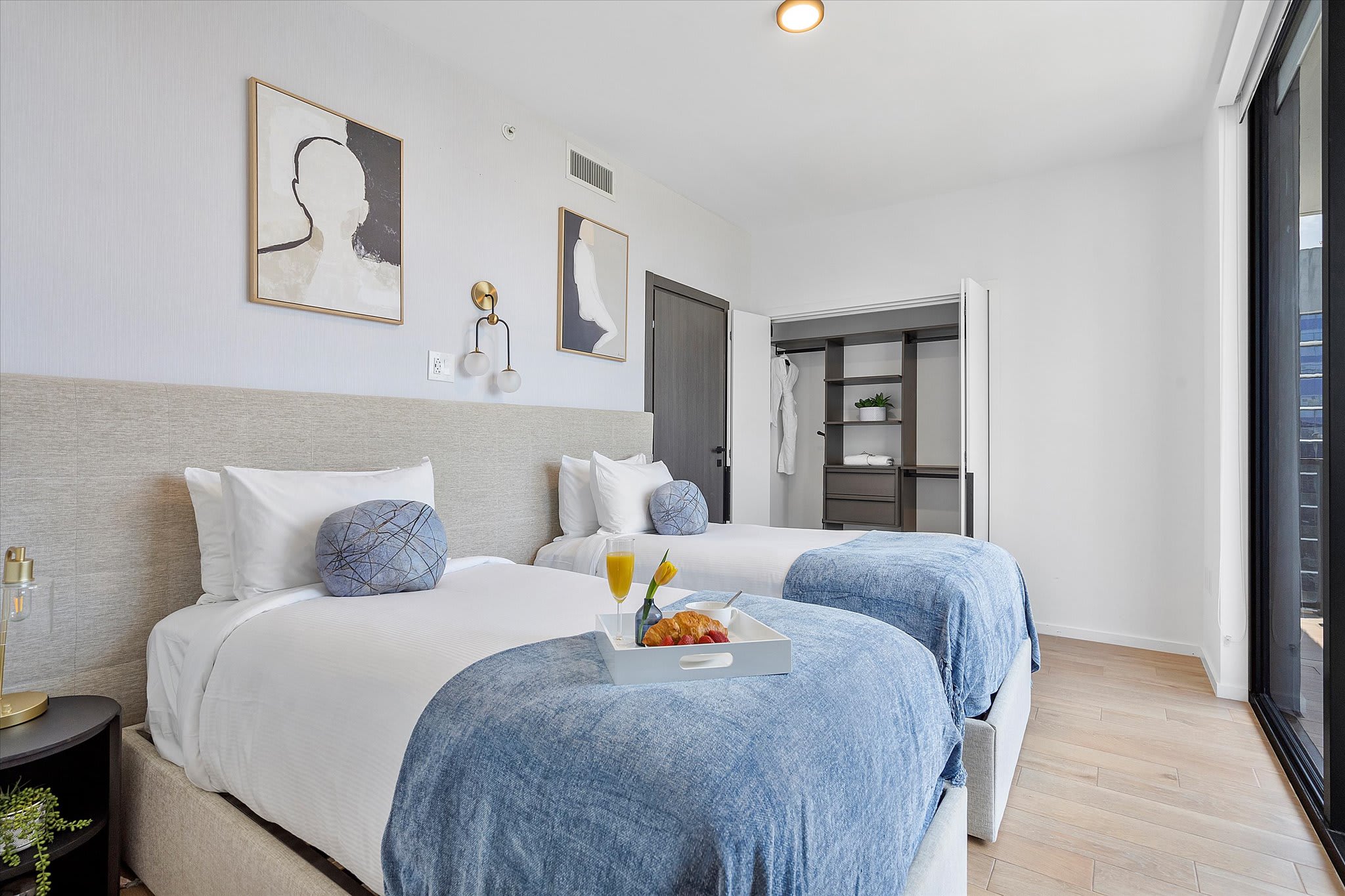 A bright bedroom featuring two twin beds with white bedding and blue throws, light wood flooring, and modern abstract art on the walls, with a breakfast tray on one bed.