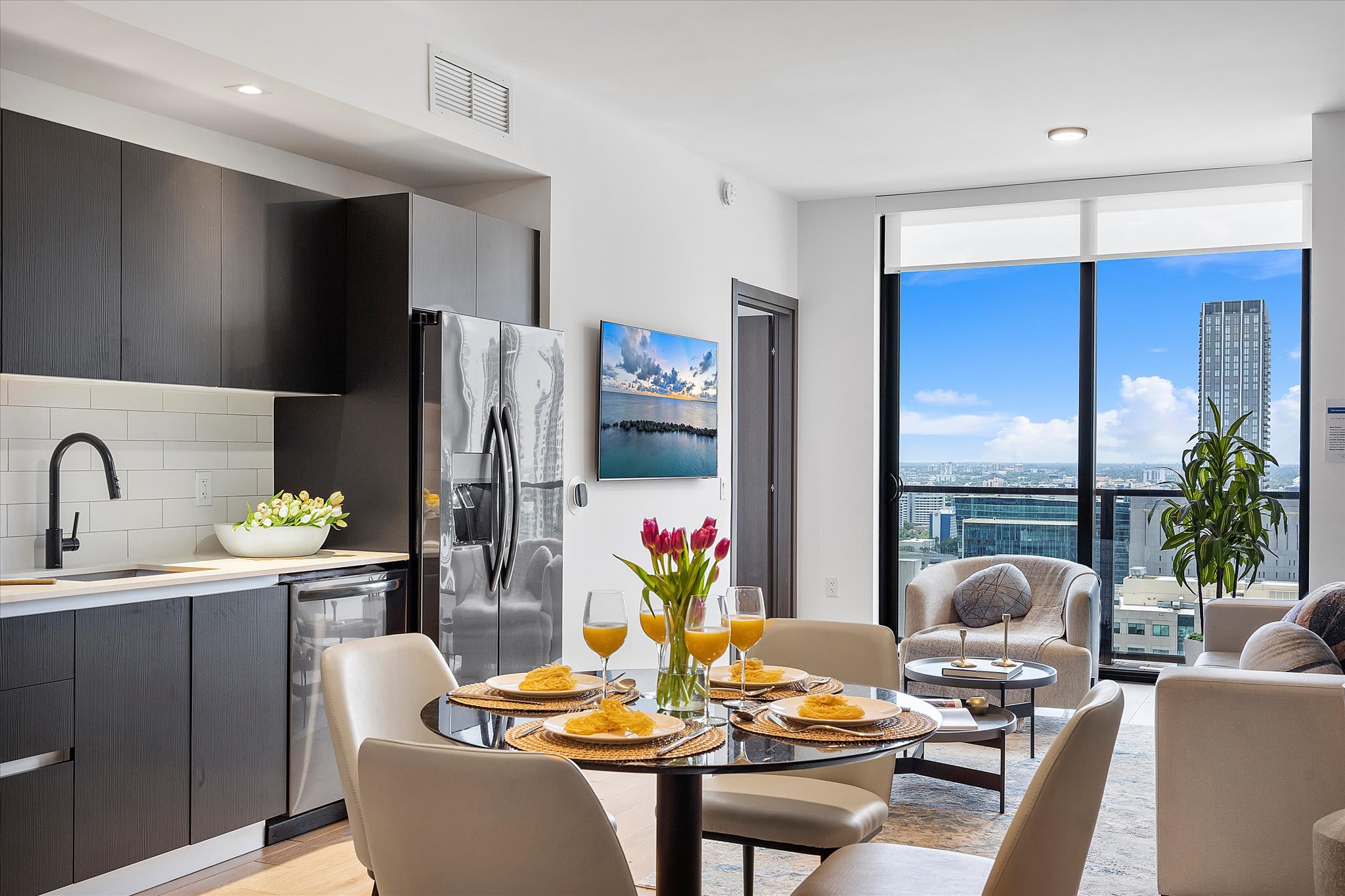 Open-concept living space featuring a modern kitchen with dark cabinetry and stainless steel appliances, a dining area with a round glass table, and a living room with a sofa and armchair. Floor-to-ceiling windows offer expansive city views, complementing the light wood-look tile floors.