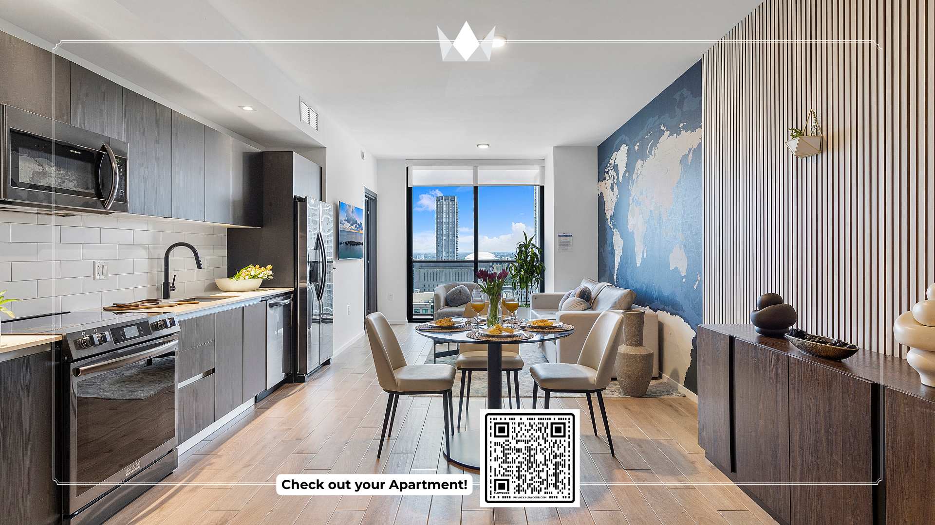 Wide shot of an open-concept living space, showcasing a modern kitchen with dark cabinetry, a dining area with a round table, and a living room with a sofa. A large blue world map accent wall adds a unique touch, and light wood-look flooring unifies the space.