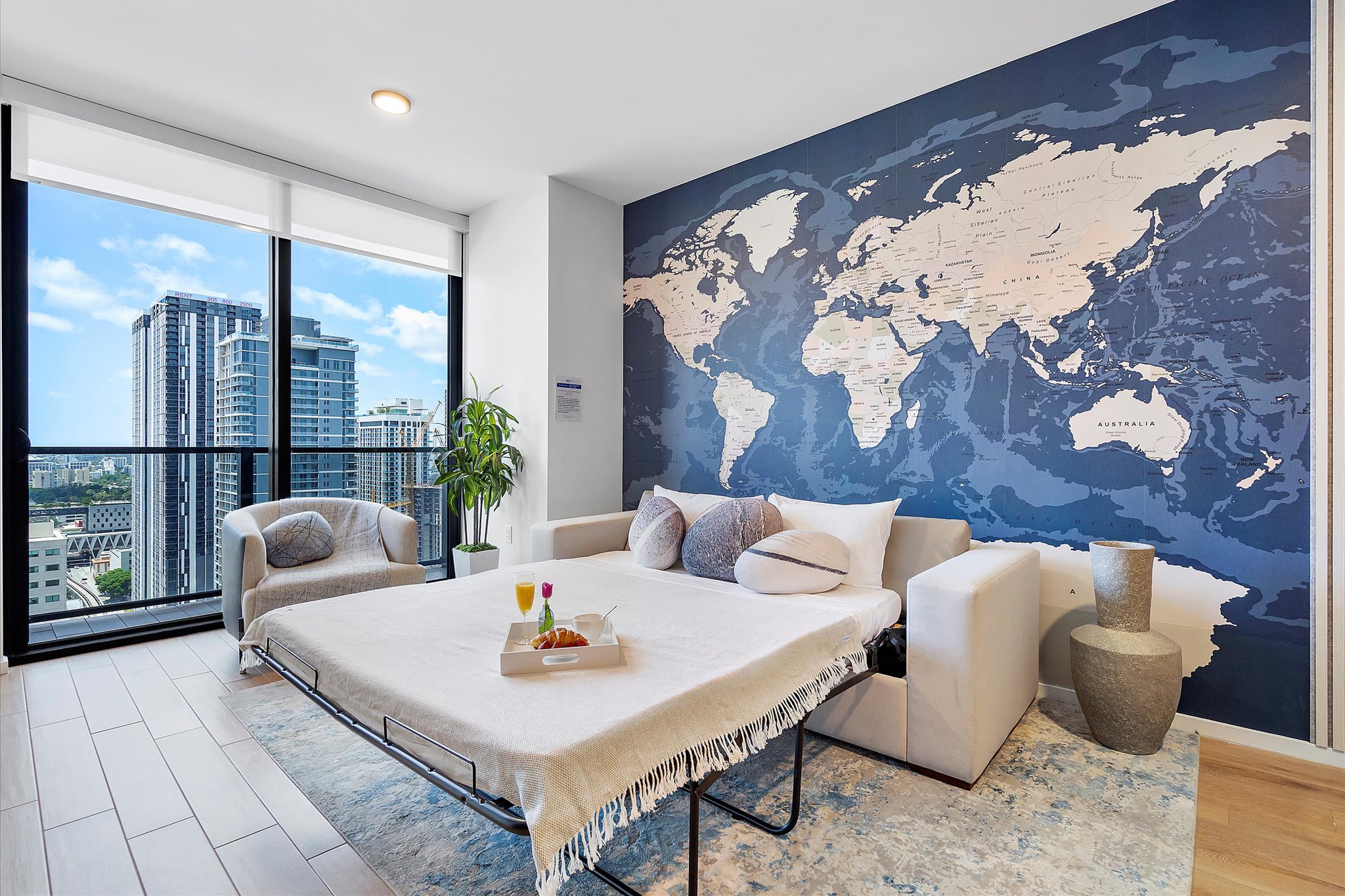 Living area transformed into a sleeping space with a pull-out sofa bed, set against a striking blue world map accent wall. Floor-to-ceiling windows offer city views, and a plush armchair provides additional seating in this bright, modern room.