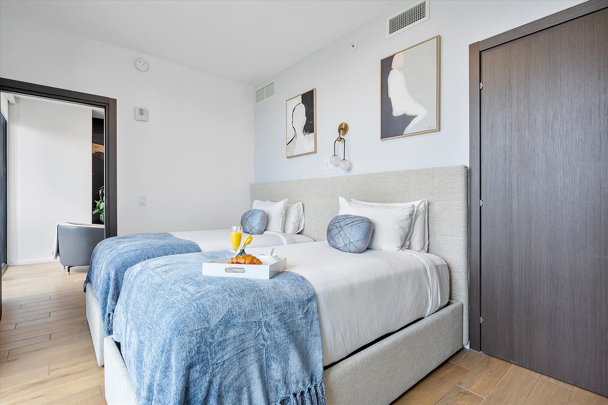A clean and modern bedroom with two twin beds, white linens, and blue accents, featuring light wood floors and abstract artwork, with a dark door leading to another room.