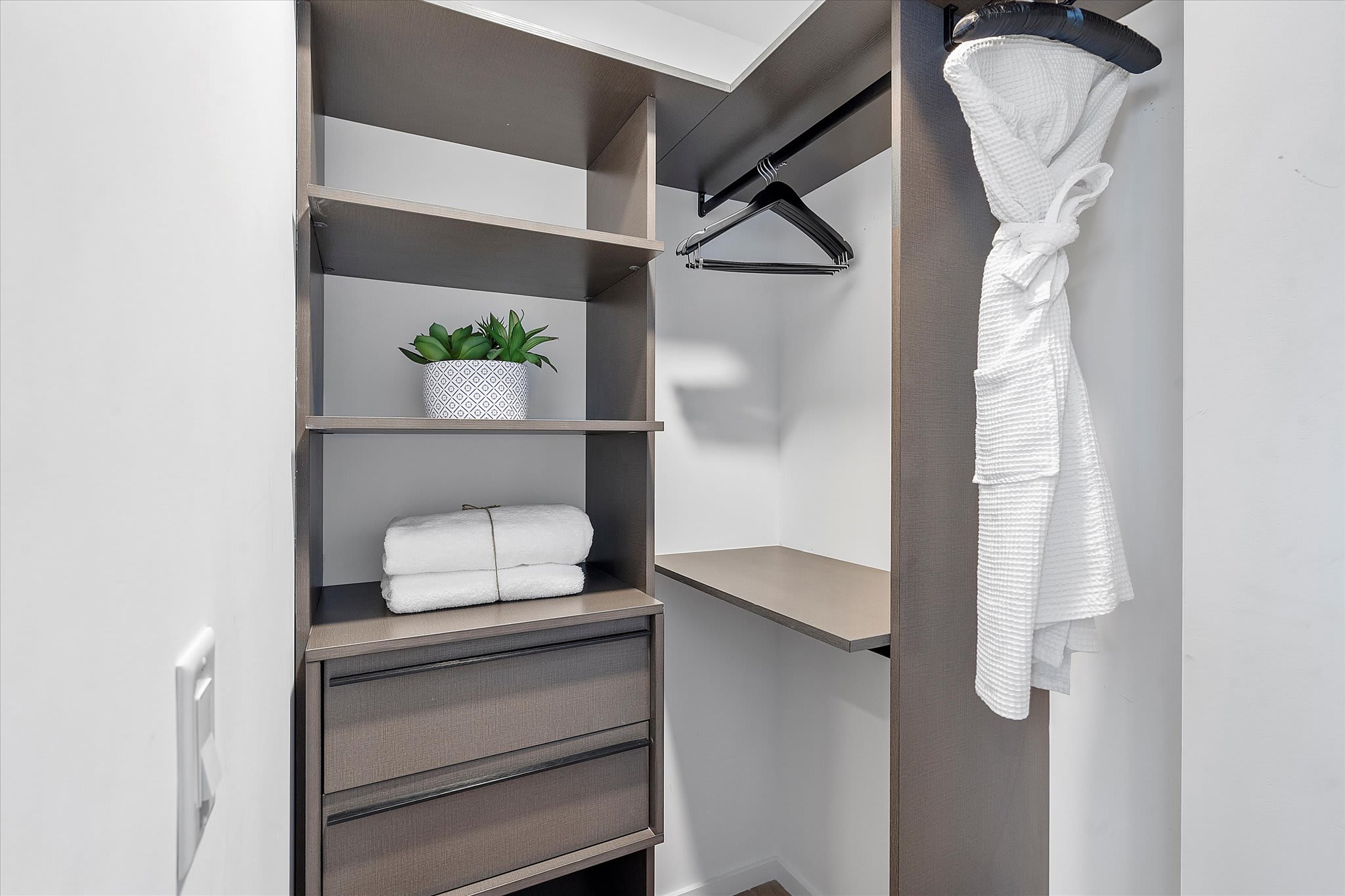 Organized walk-in closet with built-in shelving, drawers, and hanging rods. A white bathrobe and neatly folded towels are visible, along with a decorative plant.