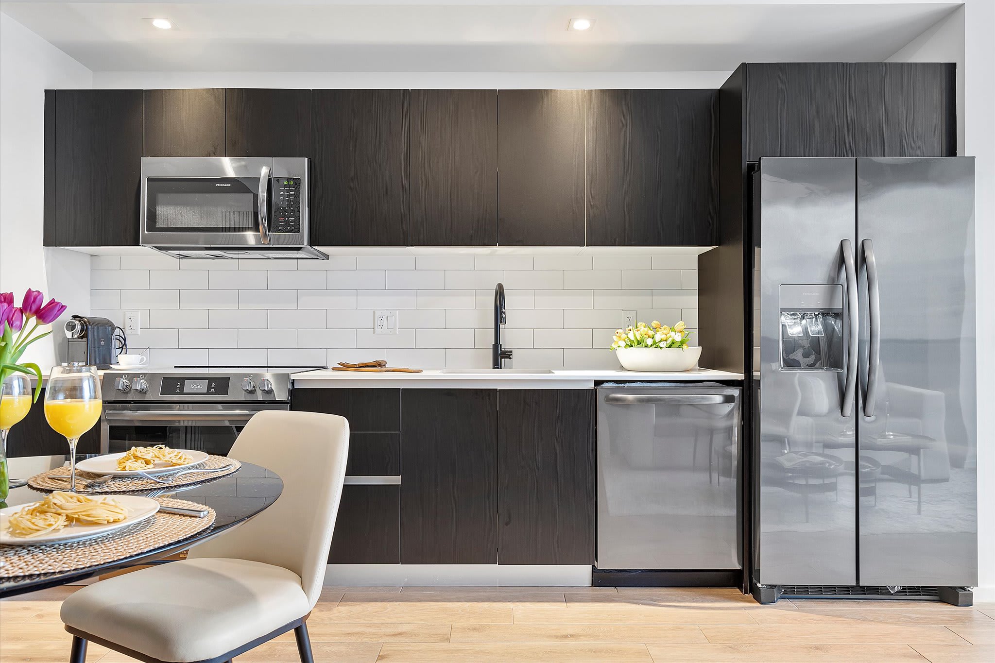 Contemporary kitchen featuring dark flat-panel cabinetry, white subway tile backsplash, and stainless steel appliances including a microwave, range, dishwasher, and French door refrigerator. A black sink with a modern faucet completes the sleek design.