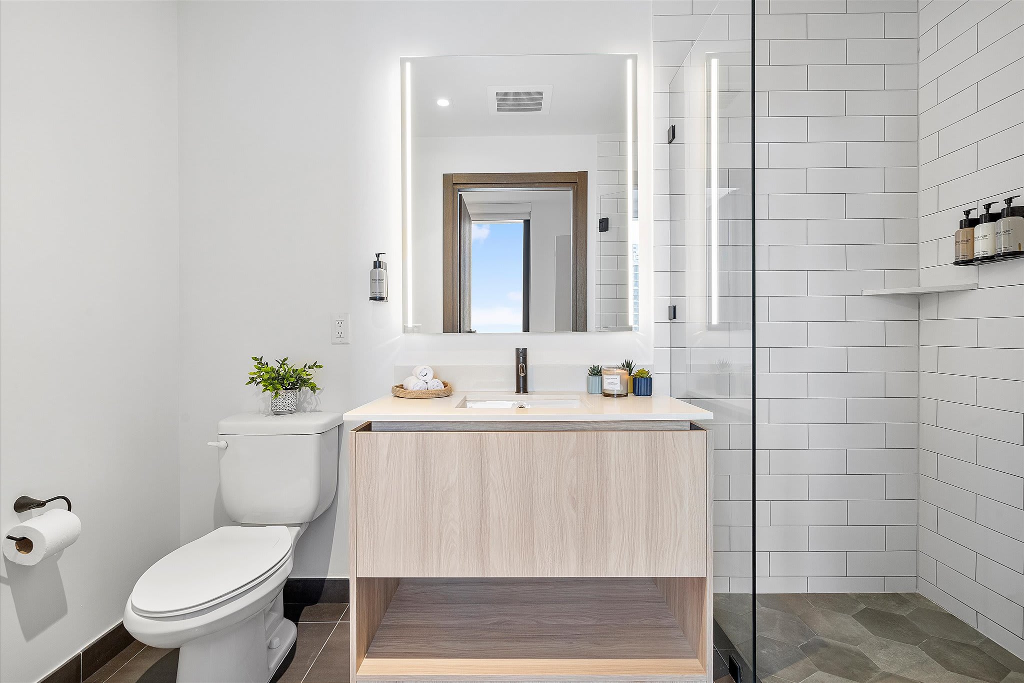 A modern bathroom with a light wood vanity, a large illuminated mirror, a white toilet, and a spacious walk-in shower with white subway tiles and dark hexagonal floor tiles.