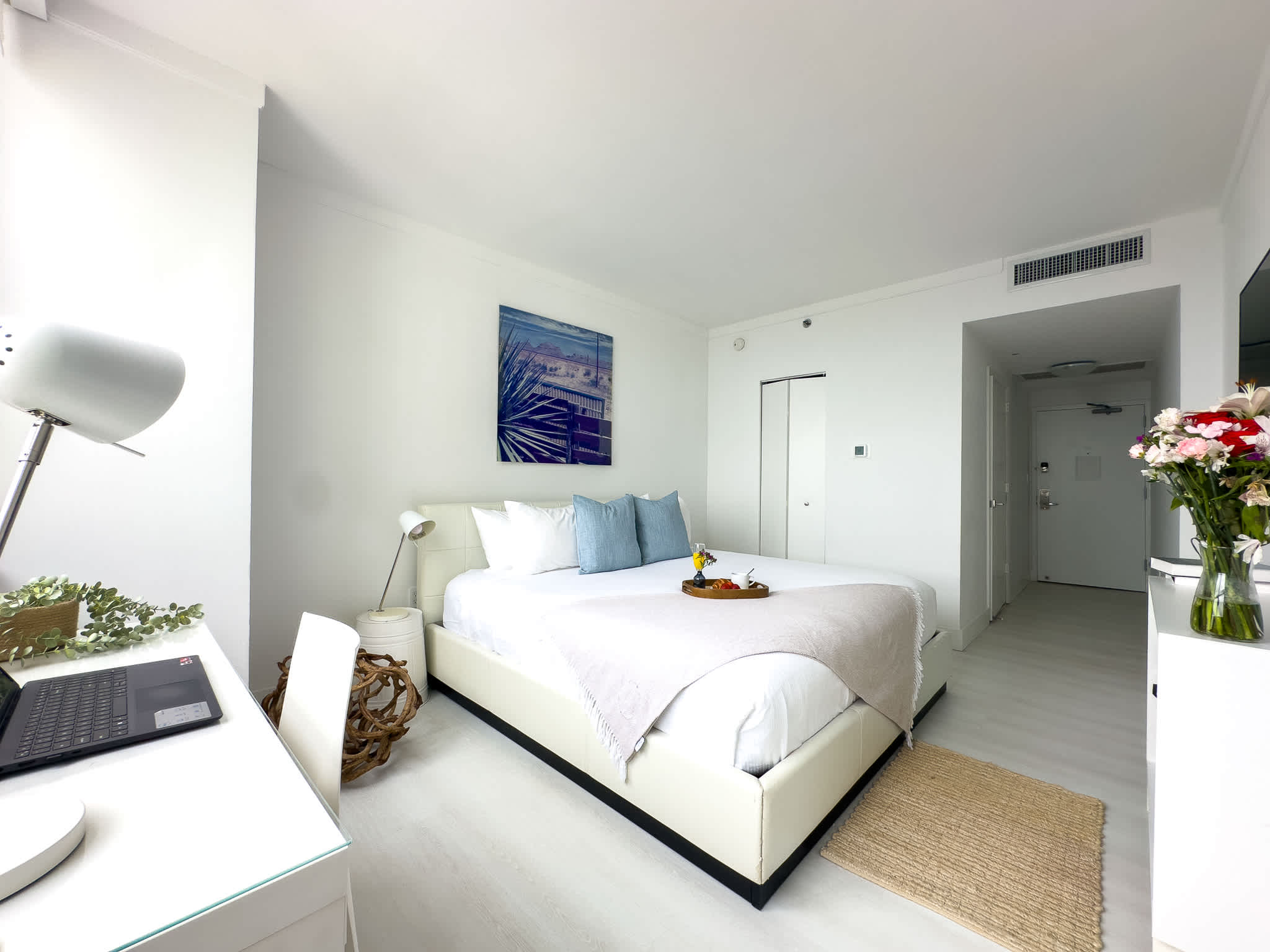 A clean, modern bedroom with a white upholstered bed, a white desk with a laptop, and a flat-screen TV on a white console. Fresh flowers add a pop of color, and light flooring complements the white walls.