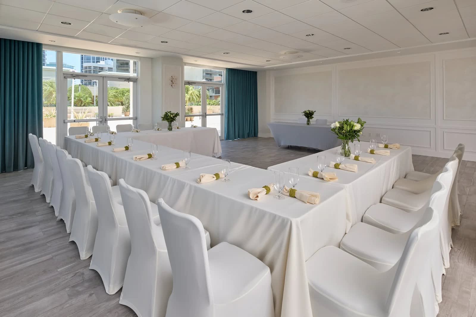 A bright conference room set up in a U-shape with white tablecloths, white chair covers, and small floral centerpieces. Large windows with blue curtains provide natural light, complementing the paneled accent wall.