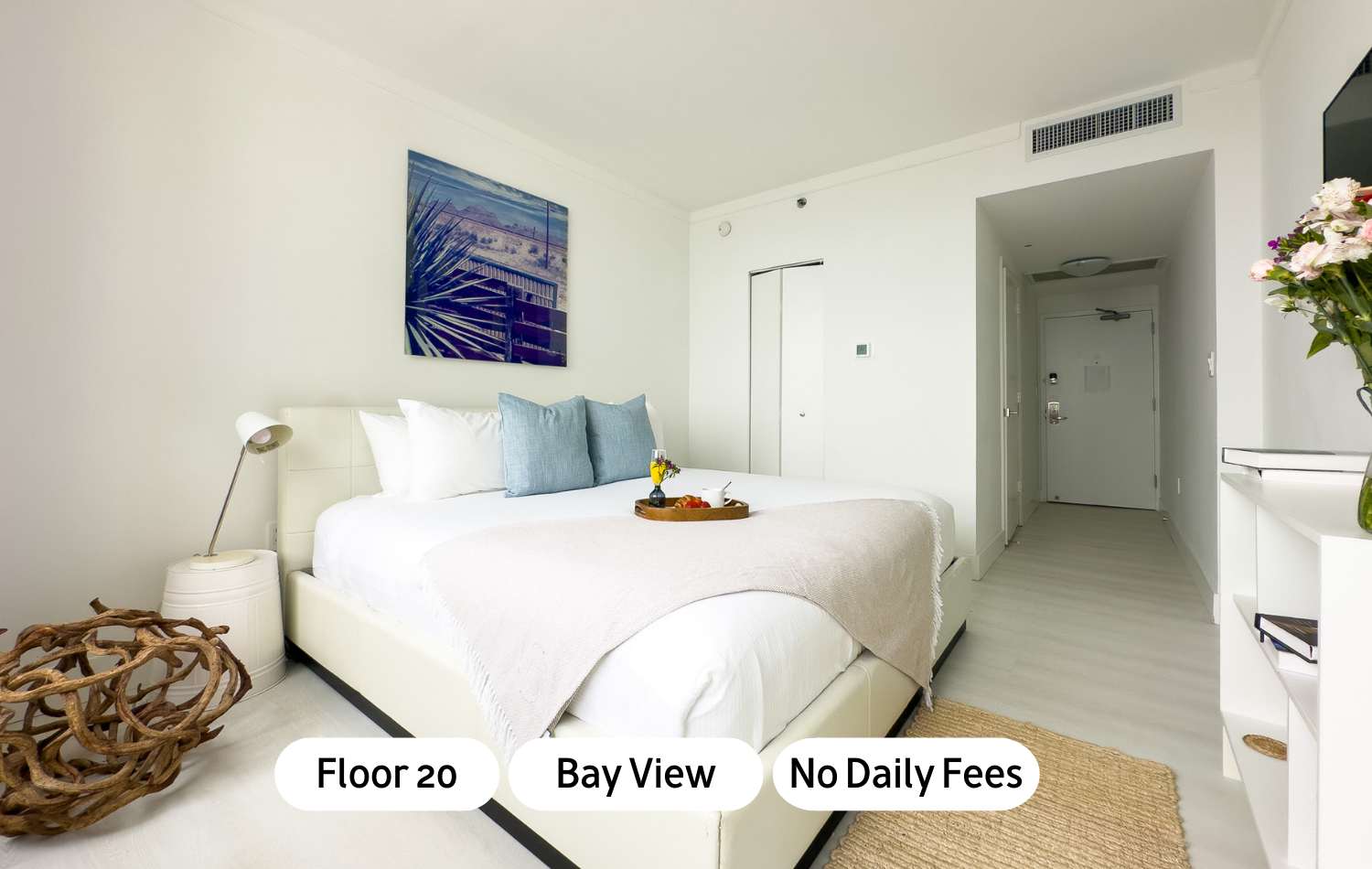 A bright, modern bedroom with a white upholstered bed, light blue pillows, and a light-colored throw blanket. A wooden tray with breakfast items sits on the bed, and a contemporary artwork hangs above the headboard.