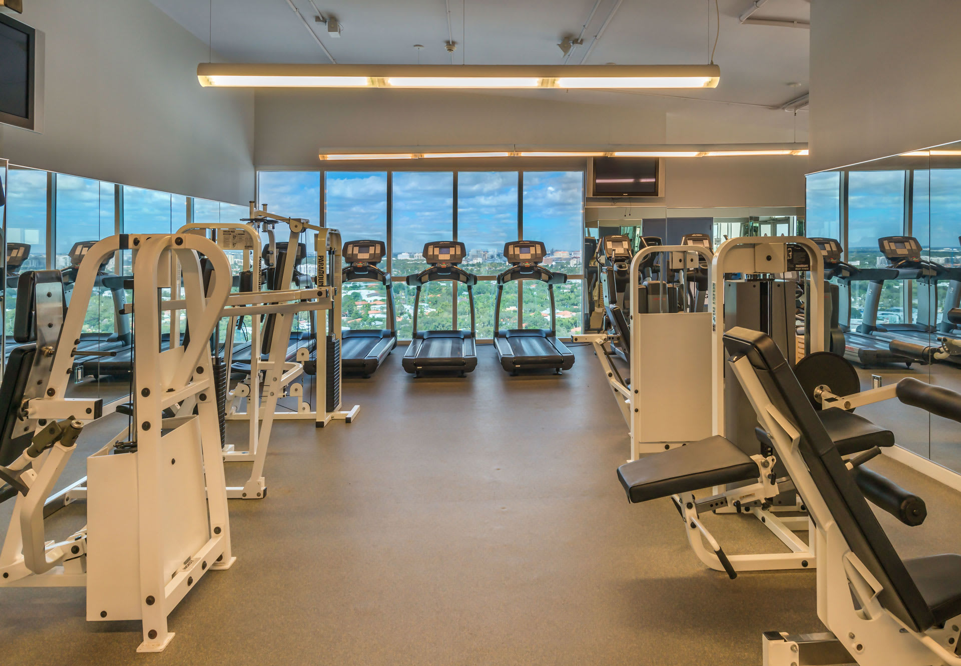 A spacious and modern gym features a variety of exercise equipment, including treadmills facing large windows with city views, and weight machines. Mirrored walls reflect the equipment, enhancing the sense of space.