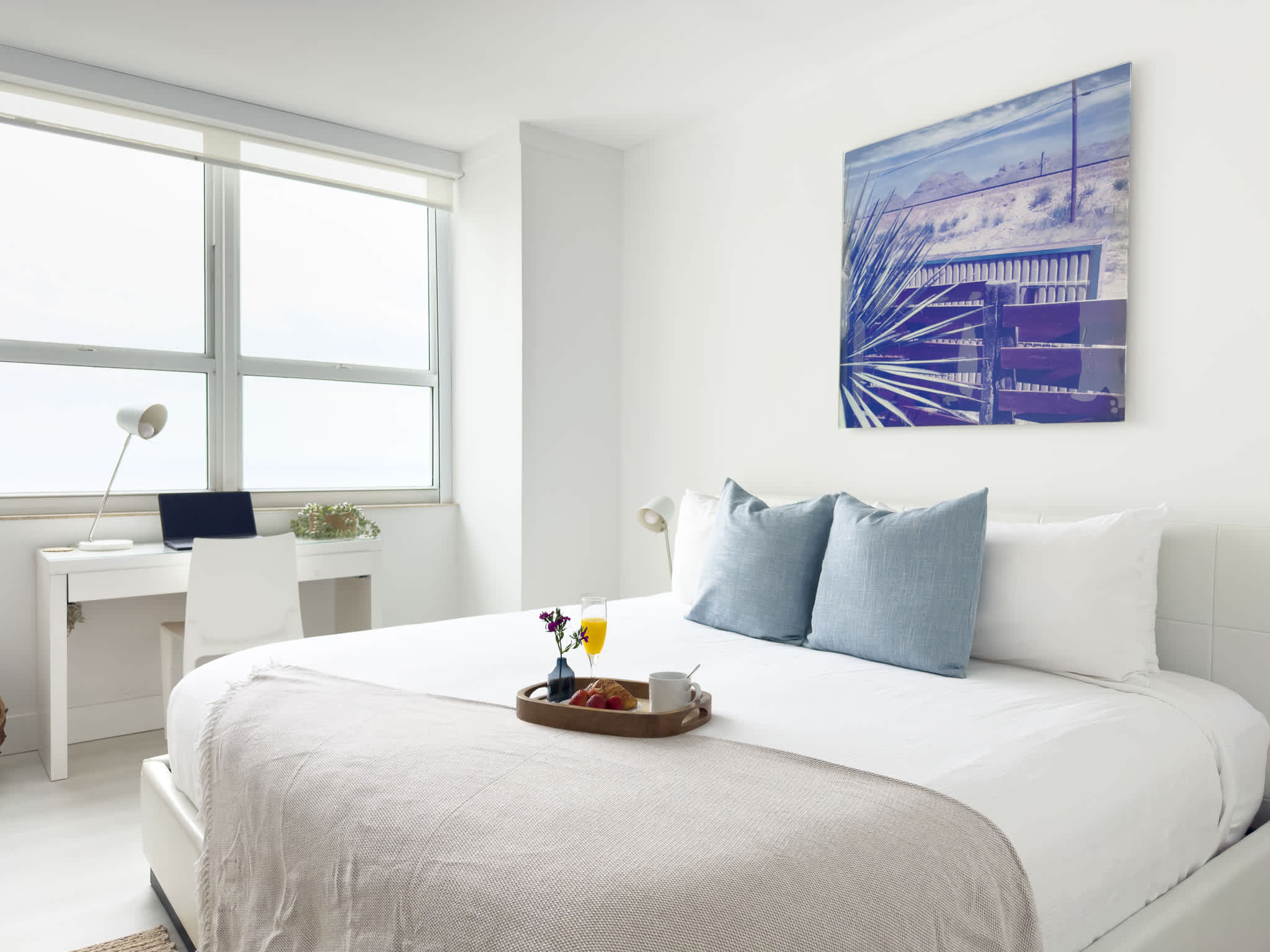 A bright bedroom featuring a white upholstered bed with light blue pillows, a modern white desk with a laptop by a large window, and a contemporary artwork above the bed.