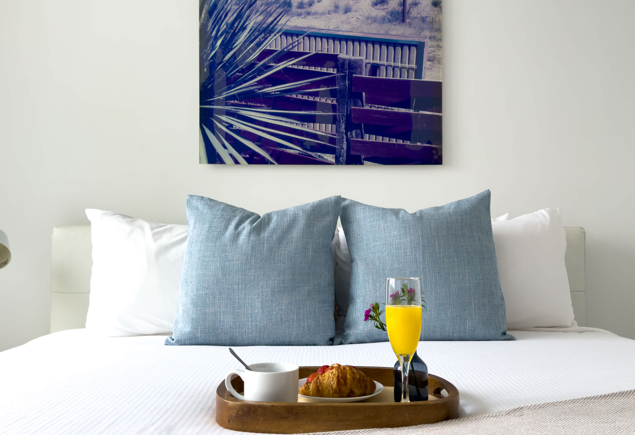 A close-up of a neatly made bed with light blue pillows, featuring a wooden tray holding a croissant, coffee, orange juice, and a small flower, set against a modern artwork on the wall.