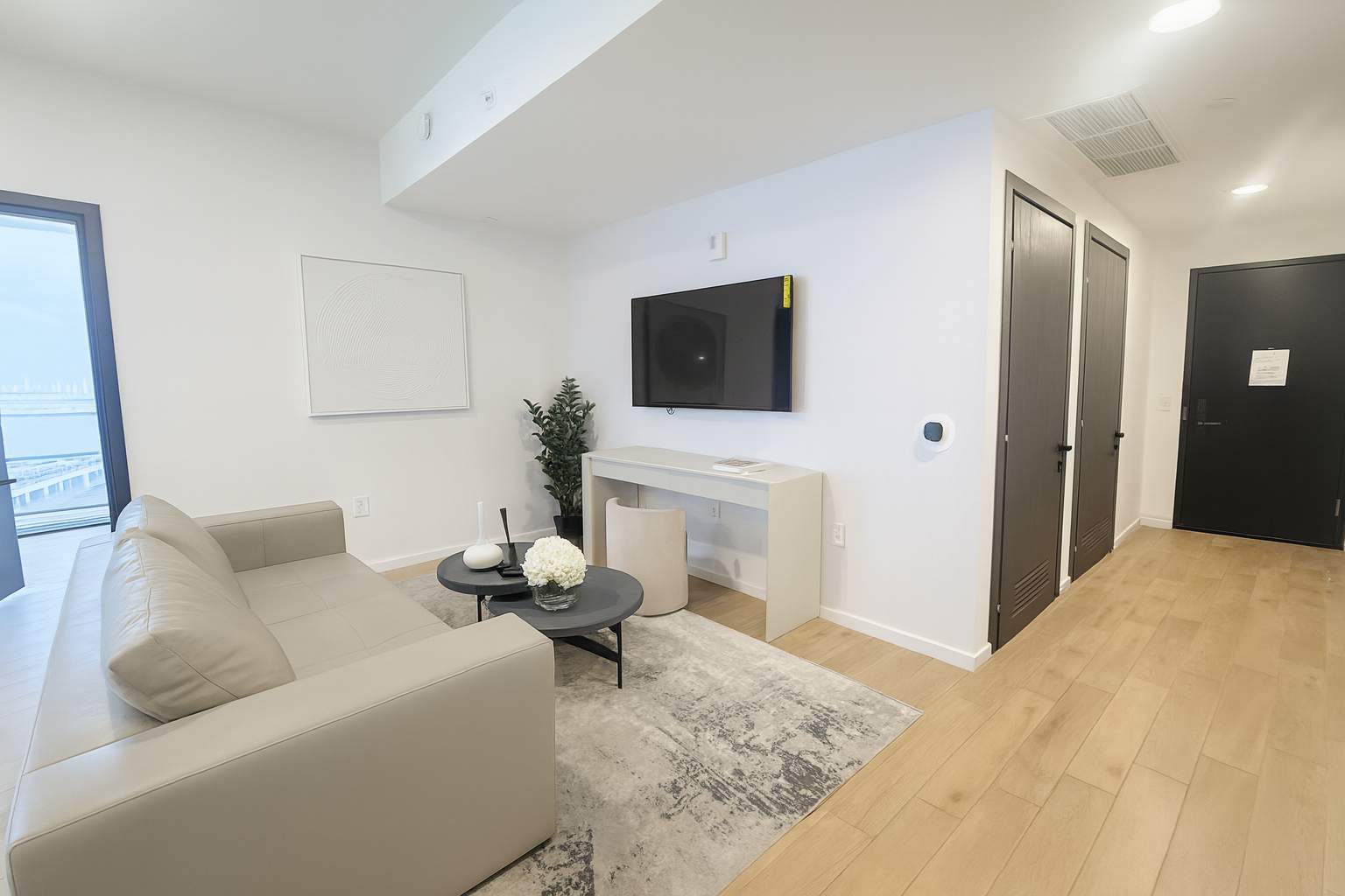 View of the living room showing a wall-mounted flat-screen TV above a console table, a beige sofa, and an area rug, with dark doors leading to other rooms or closets.