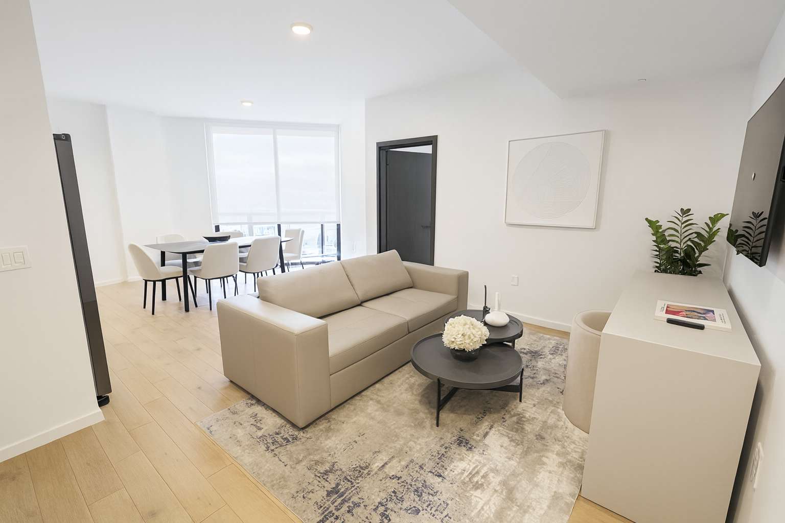 Contemporary living room furnished with a beige sofa, two round coffee tables, and a patterned area rug, with a dining area visible in the background and light hardwood floors throughout.